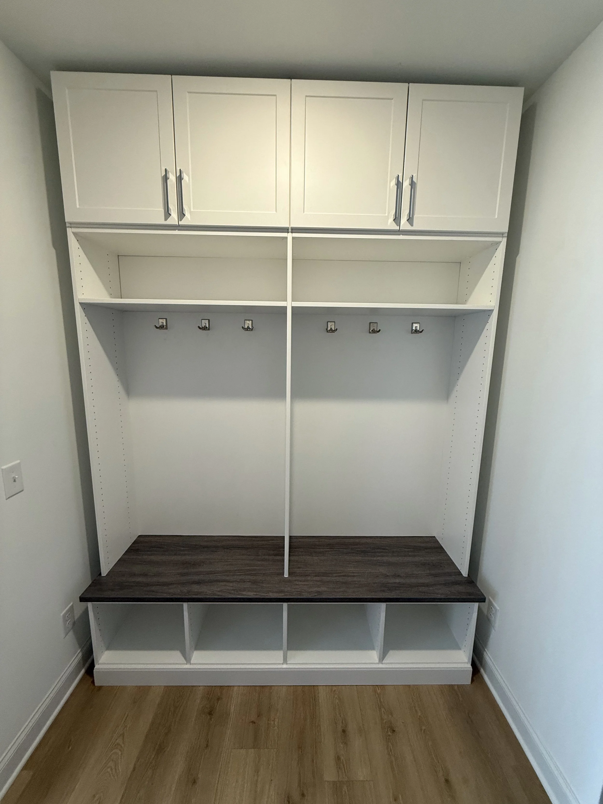 Empty white closet with cabinets, hooks, and cubbies, with a dark wood bench and a wooden floor.