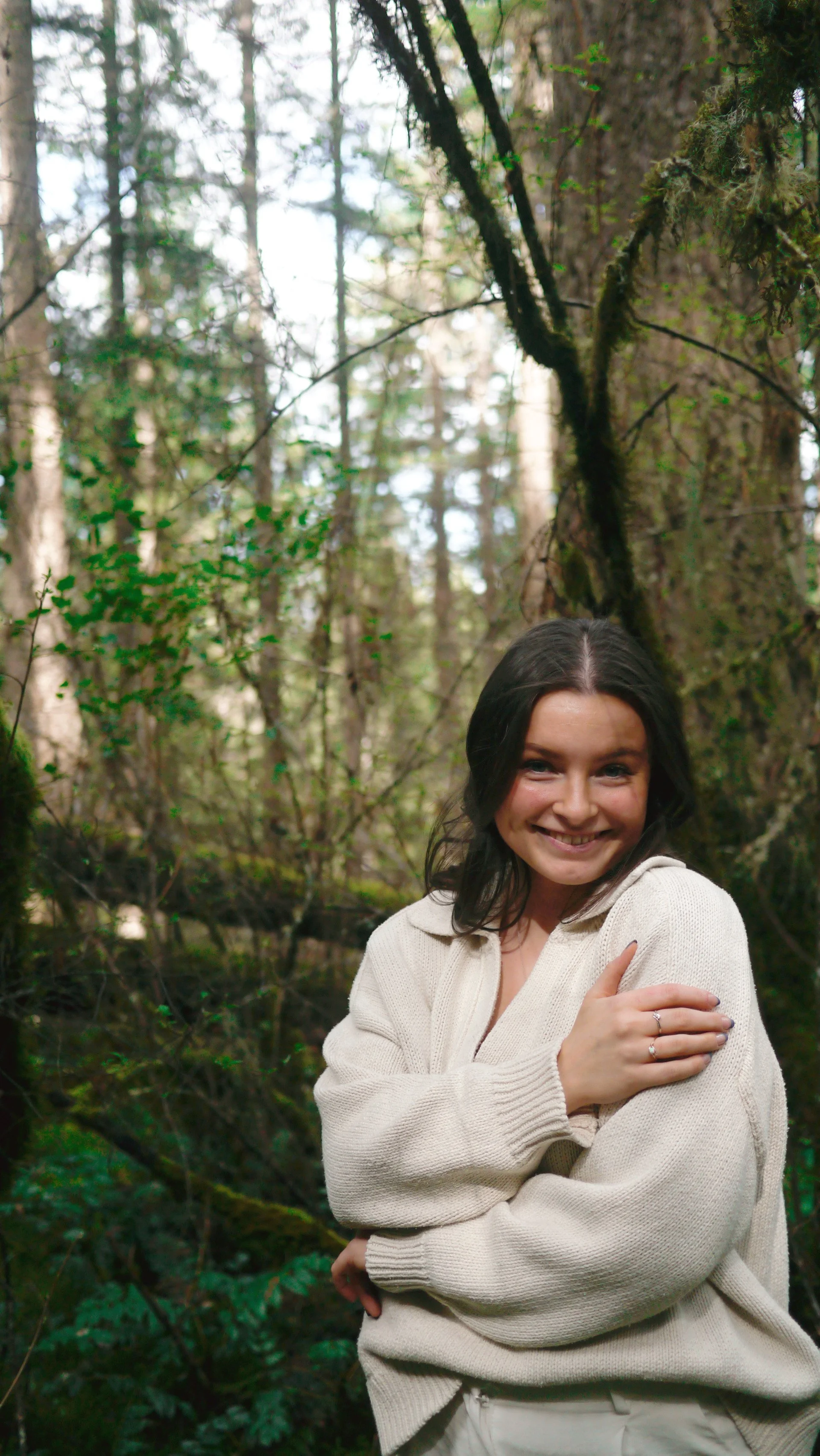 Nuala the doula, birth and postpartum doula in Victoria, BC. Ember and Earth Doula. A smiling woman with dark hair and light skin, wearing a cream-colored sweater, standing in a forest with trees and green foliage.