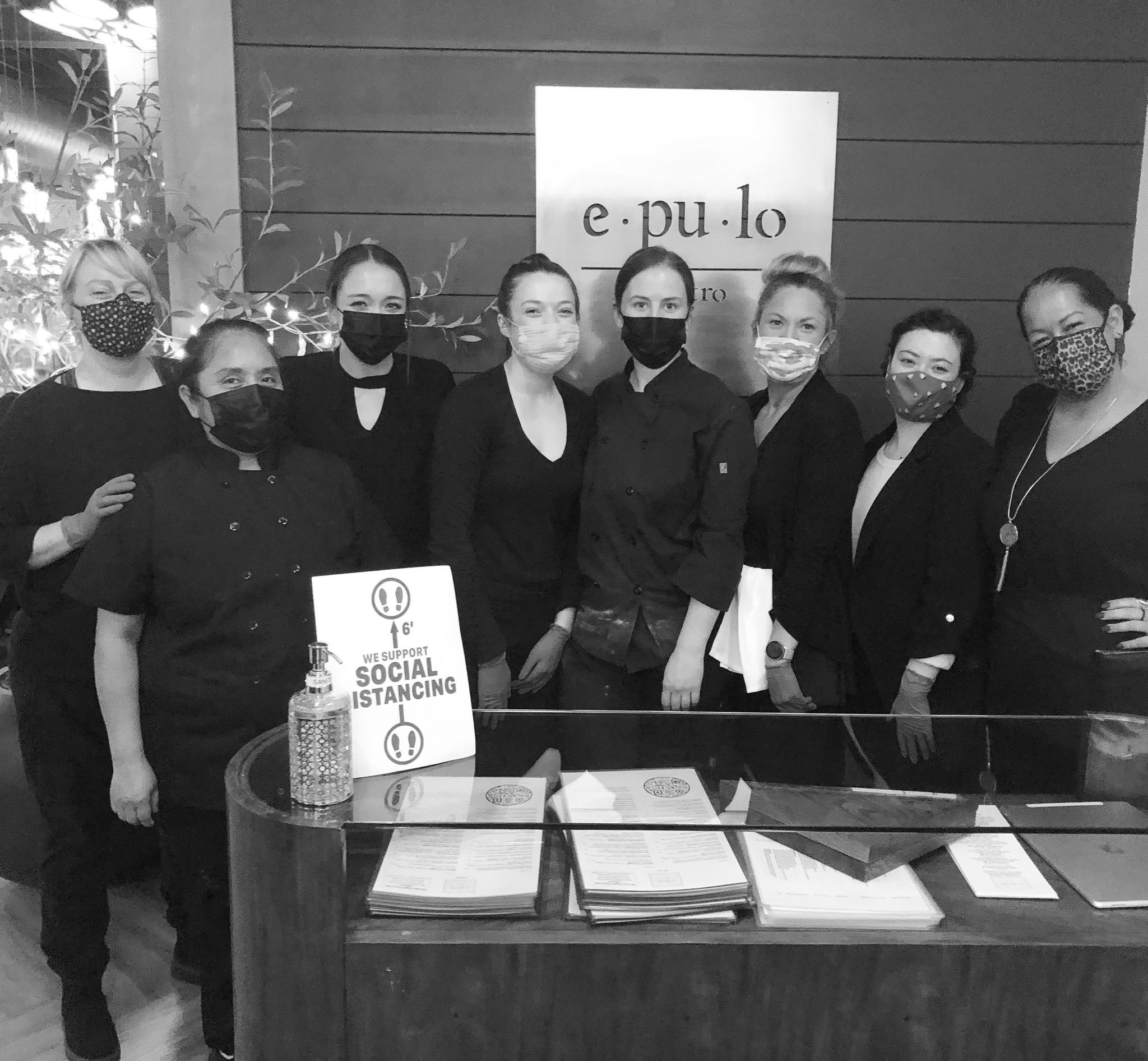 Group of women working at a restaurant or cafe, all wearing masks, standing behind a reception desk with menus and a sign indicating social distancing.