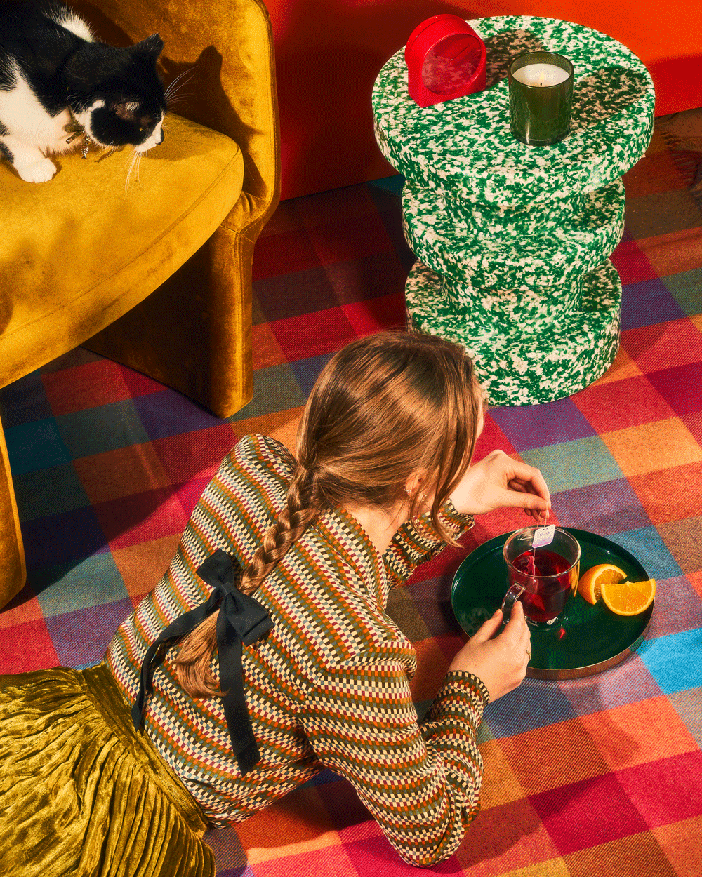 Woman lying on colorful checkered carpet, holding a cup with tea bag label, near a green terrazzo side table with candle, and a black and white cat on mustard sofa.