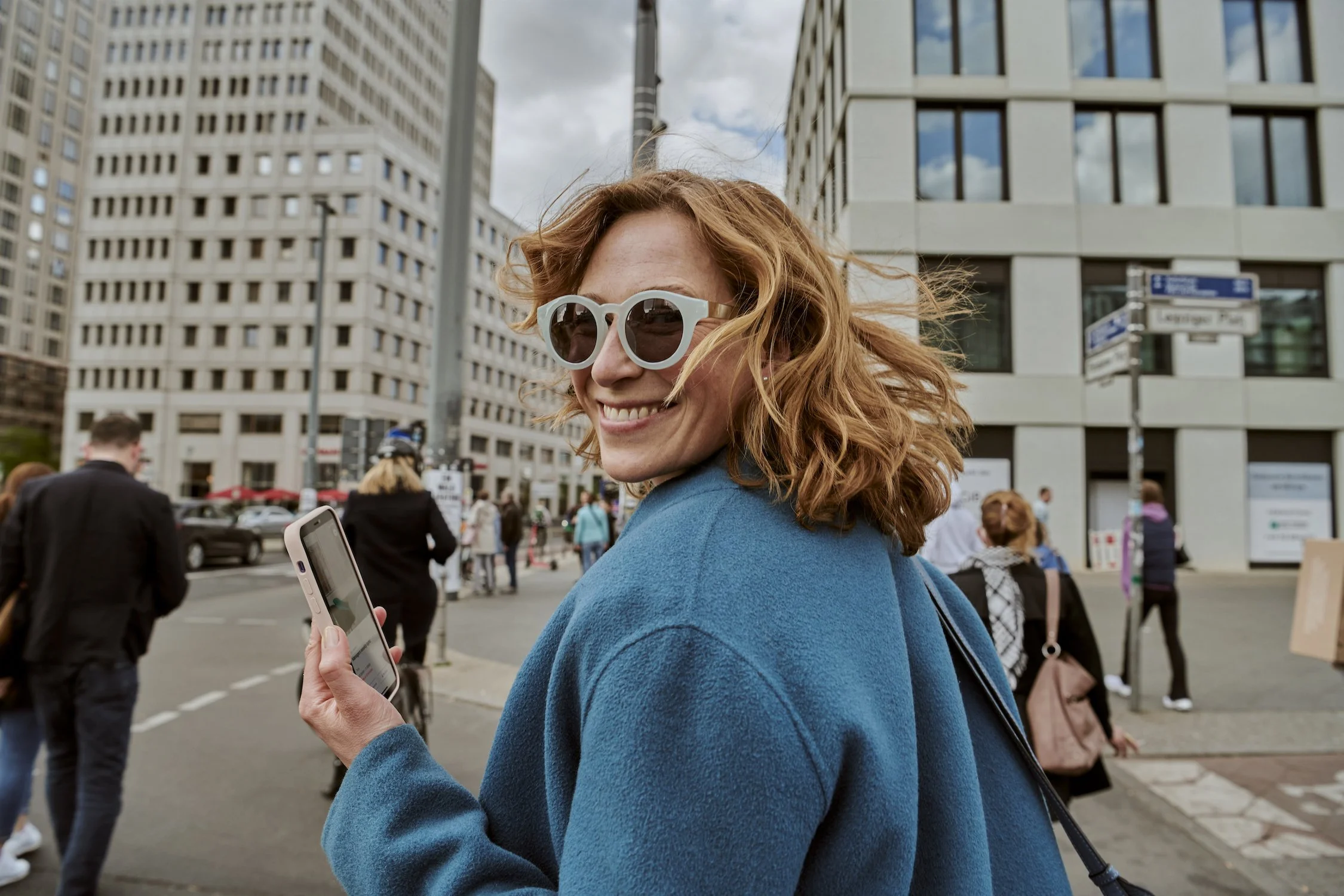 Woman in blue coat and sunglasses smiling and holding a smartphone on a city street, with modern buildings in the background.