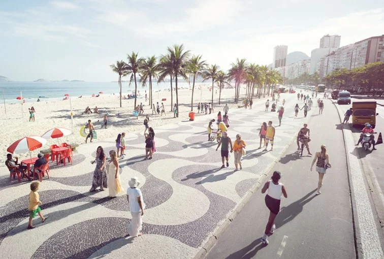 People walking and jogging along a patterned sidewalk by a beach with palm trees, red umbrellas, and distant buildings.