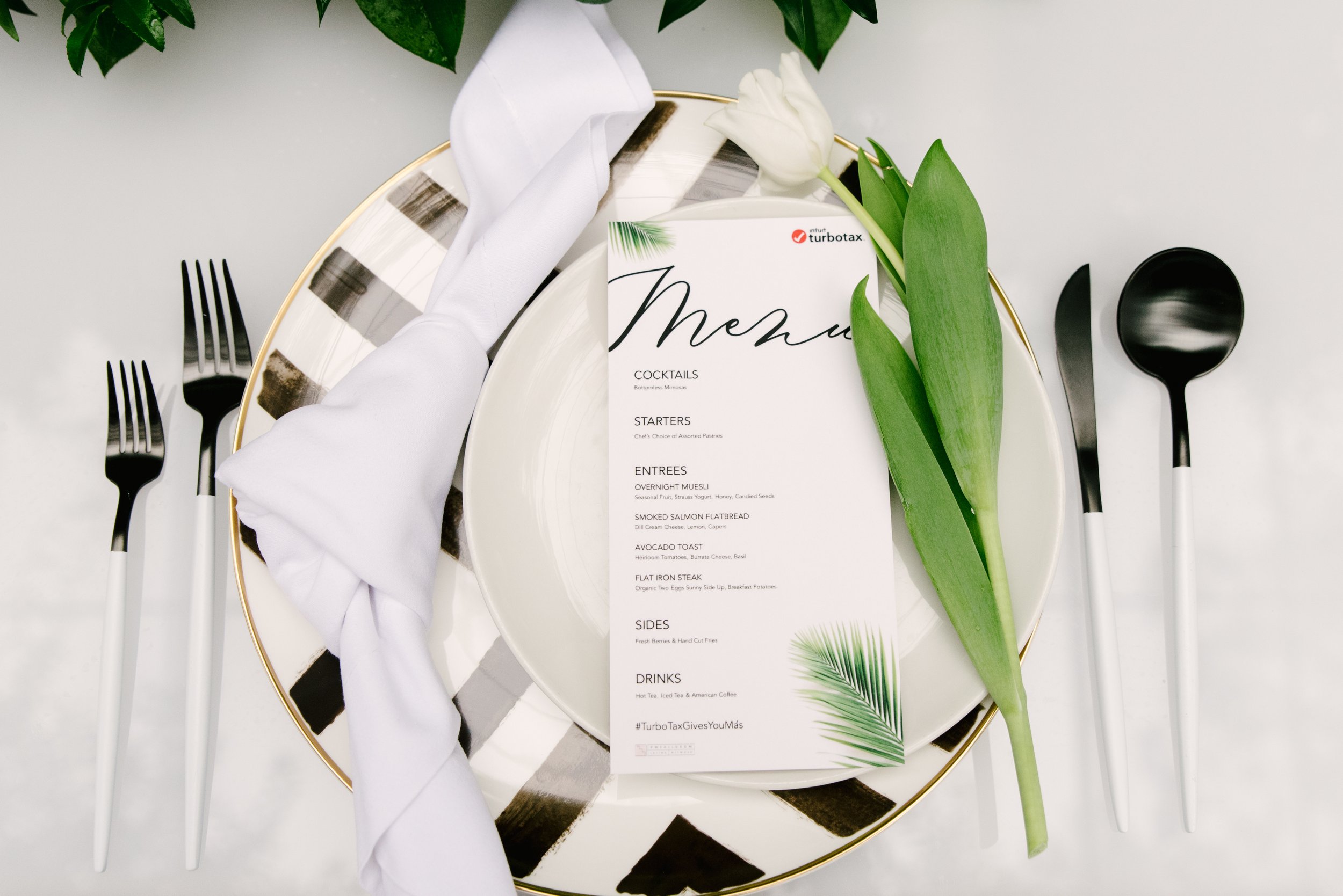 A table setting with a white plate, a white napkin, silverware, a white tulip flower, and a menu card, decorated with green leaves.