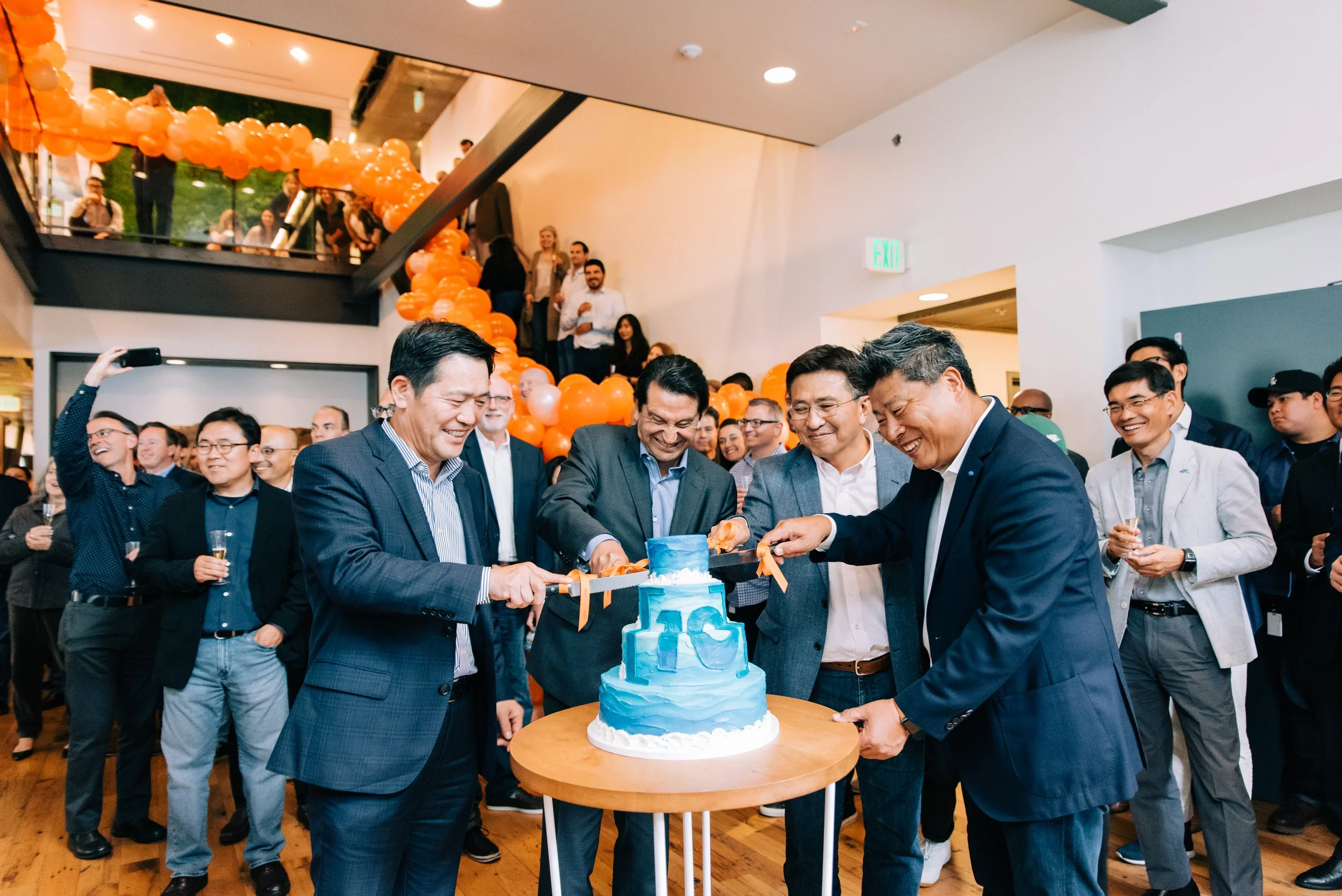 Corporate grand opening event in Irvine, featuring executives cutting a celebratory blue cake while guests cheer beneath a dramatic balloon installation, captured by Tiffany Perry Photography and Photo Booth Squared.