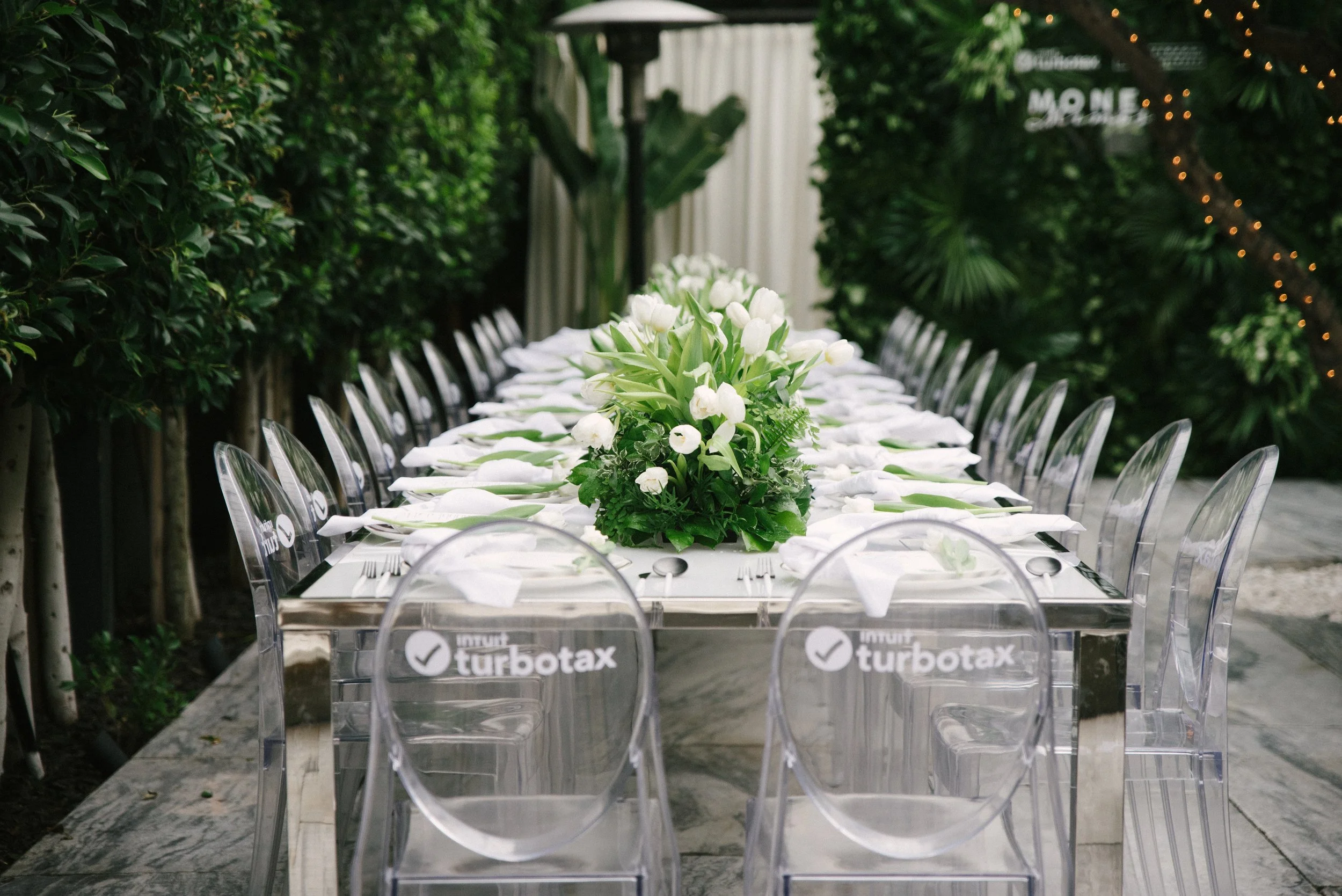  – Elegant corporate dinner event in Los Angeles, featuring a sleek modern tablescape with branded ghost chairs, lush white floral centerpieces, and a greenery-filled outdoor setting, captured by Tiffany Perry Photography and Photo Booth Squared.