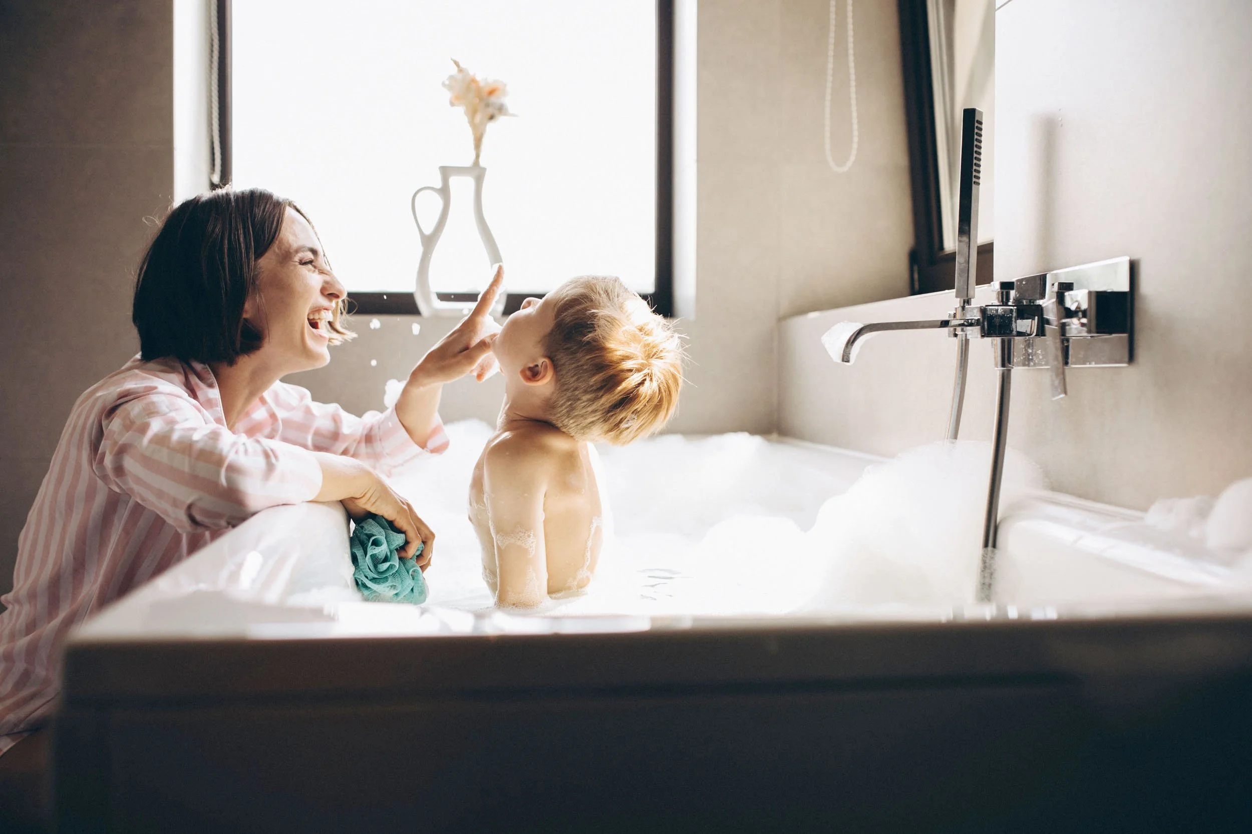 A woman and a young boy sharing a joyful moment in a bubble-filled bathtub, with the woman smiling and playfully touching the boy's nose, while a window with bright light is in the background.