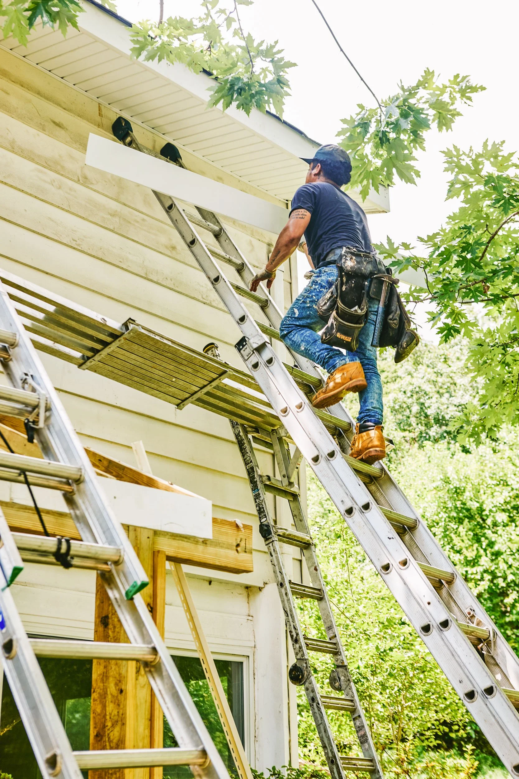A worker in blue jeans, dark shirt, boots, and a cap is climbing a ladder to perform repair on the gutters of a house, with trees and greenery around.
