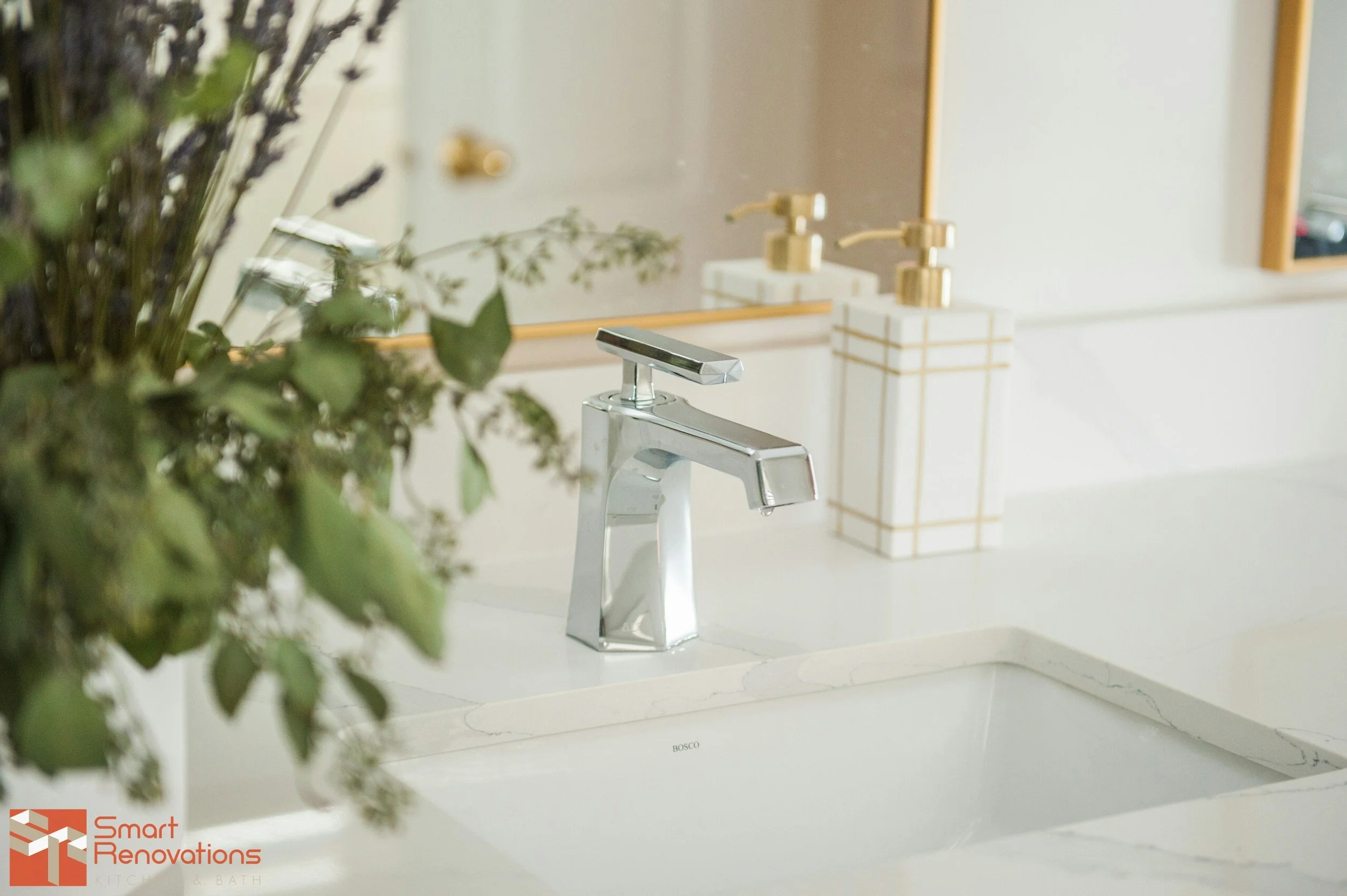 A bathroom sink with a modern chrome faucet, a mirror, and a soap dispenser with a gold pump. There is a bouquet of fresh greenery and lavenders on the left side of the image. The countertop is white with a marble-like pattern, and the background shows a mirror and a white wall.