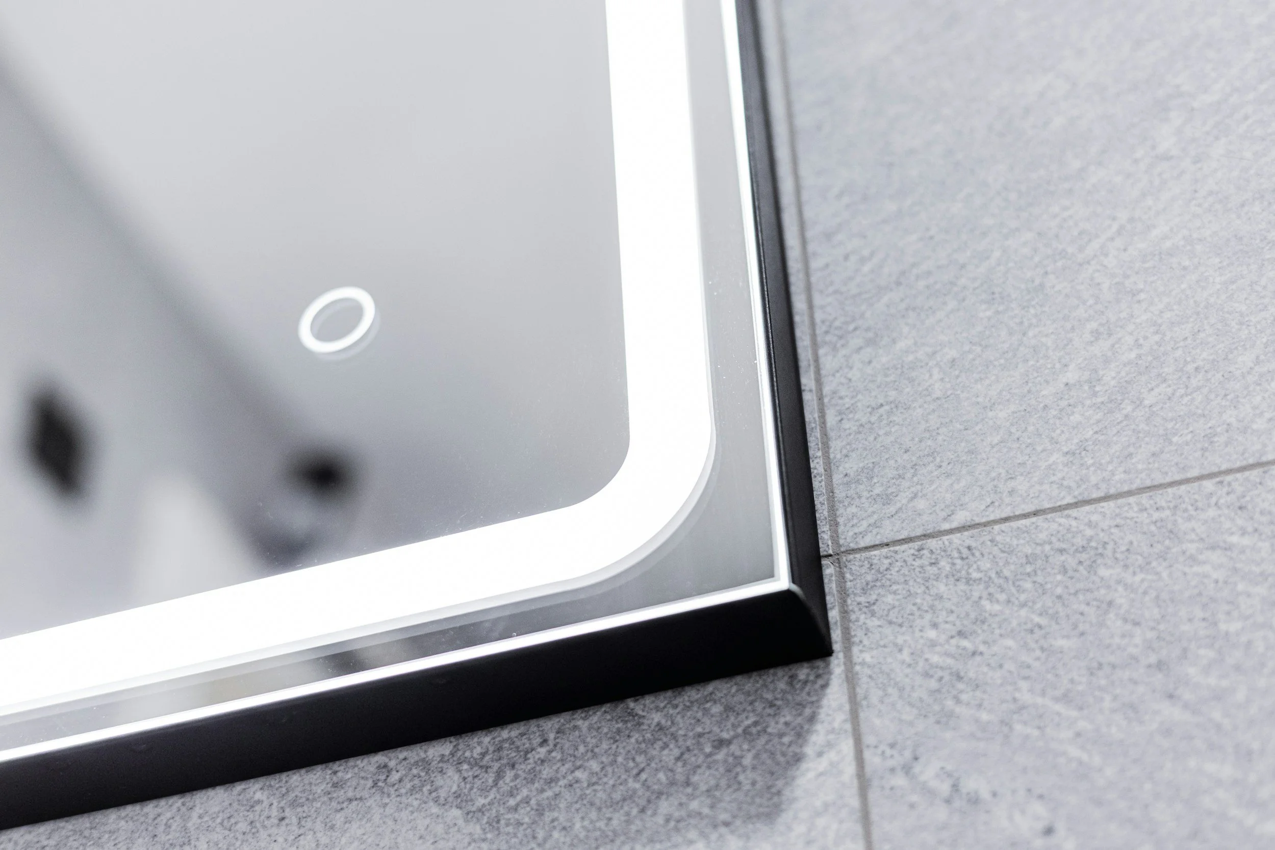 Close-up of a corner of a mirror with a black frame on a gray tiled wall.
