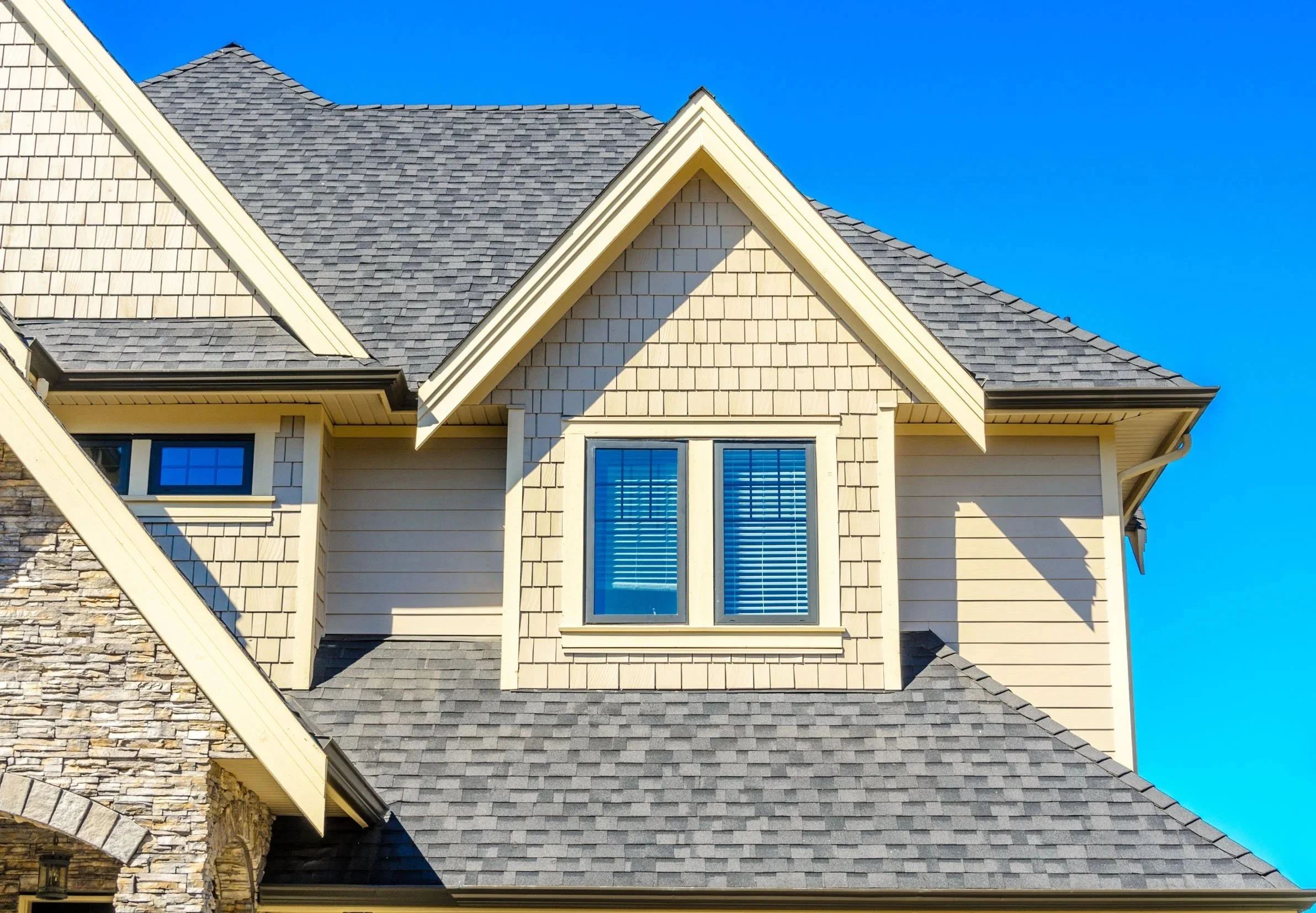 Close-up of the upper floor of a house with beige siding, stone accents, and a gray shingled roof under a clear blue sky.