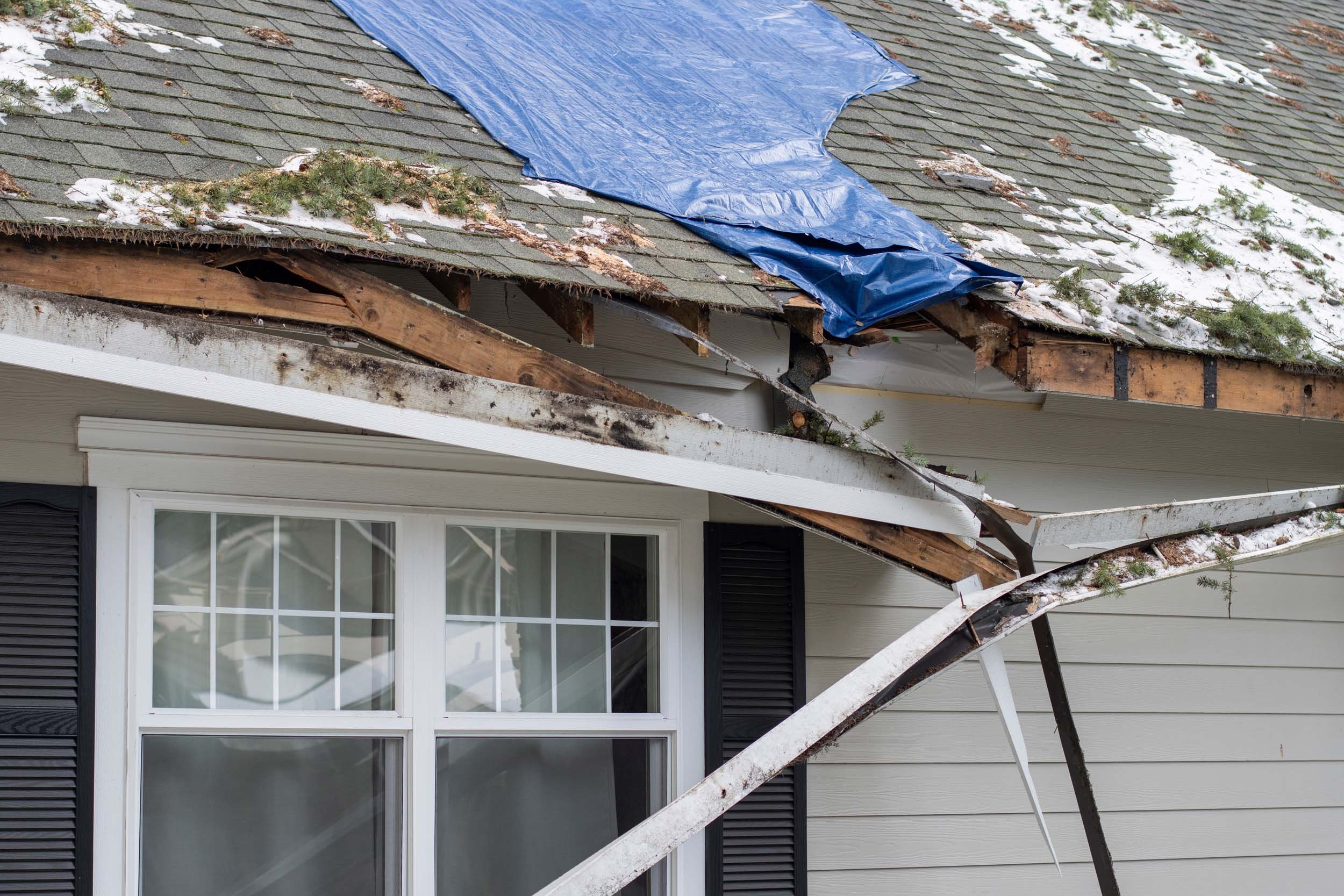 Damaged house roof with missing shingles, broken rafters, and debris, including a blue tarp covering part of the damaged roof.