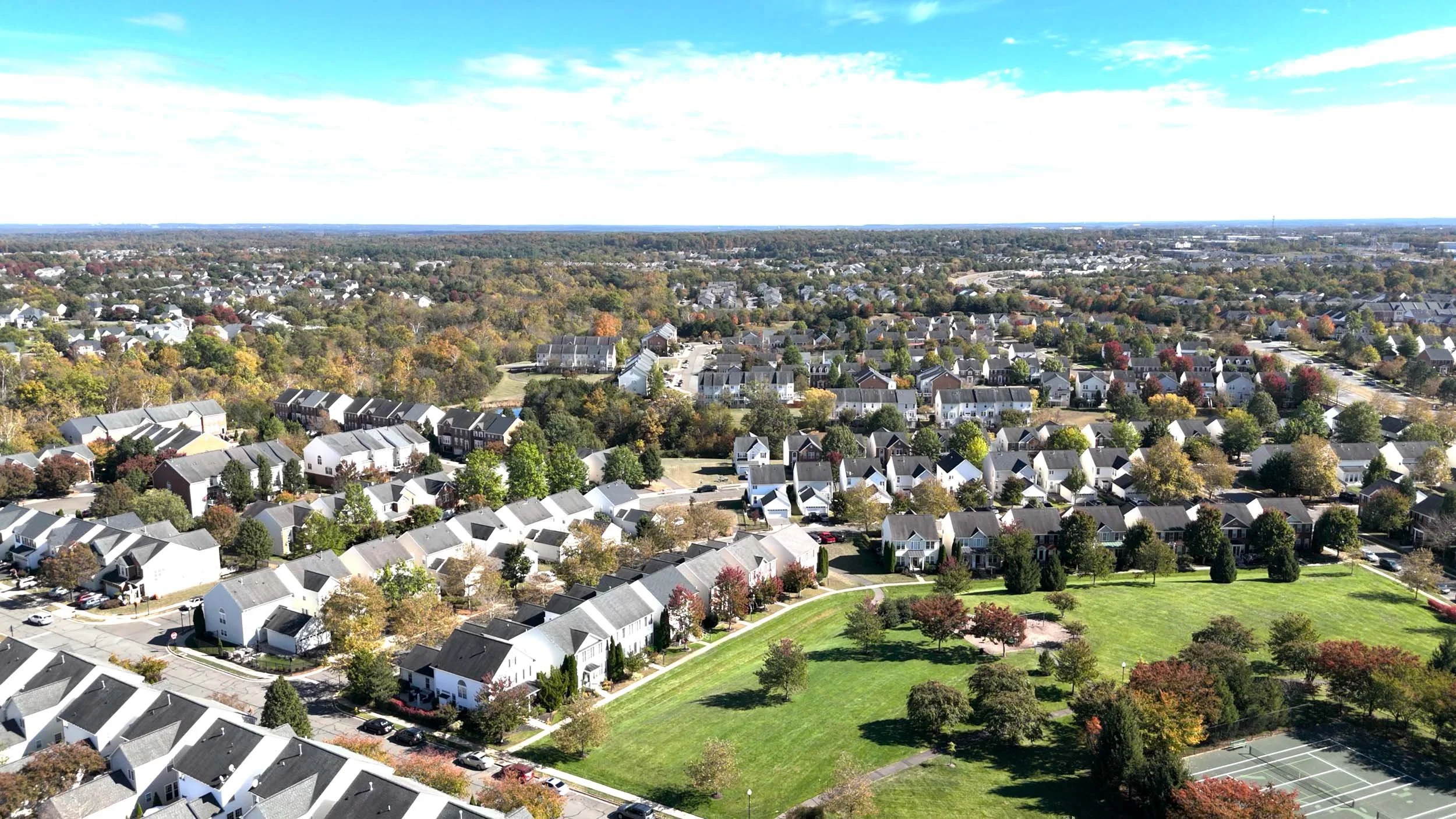 An aerial view of a suburban neighborhood with rows of houses, streets, trees, and a large grassy park under a partly cloudy sky.