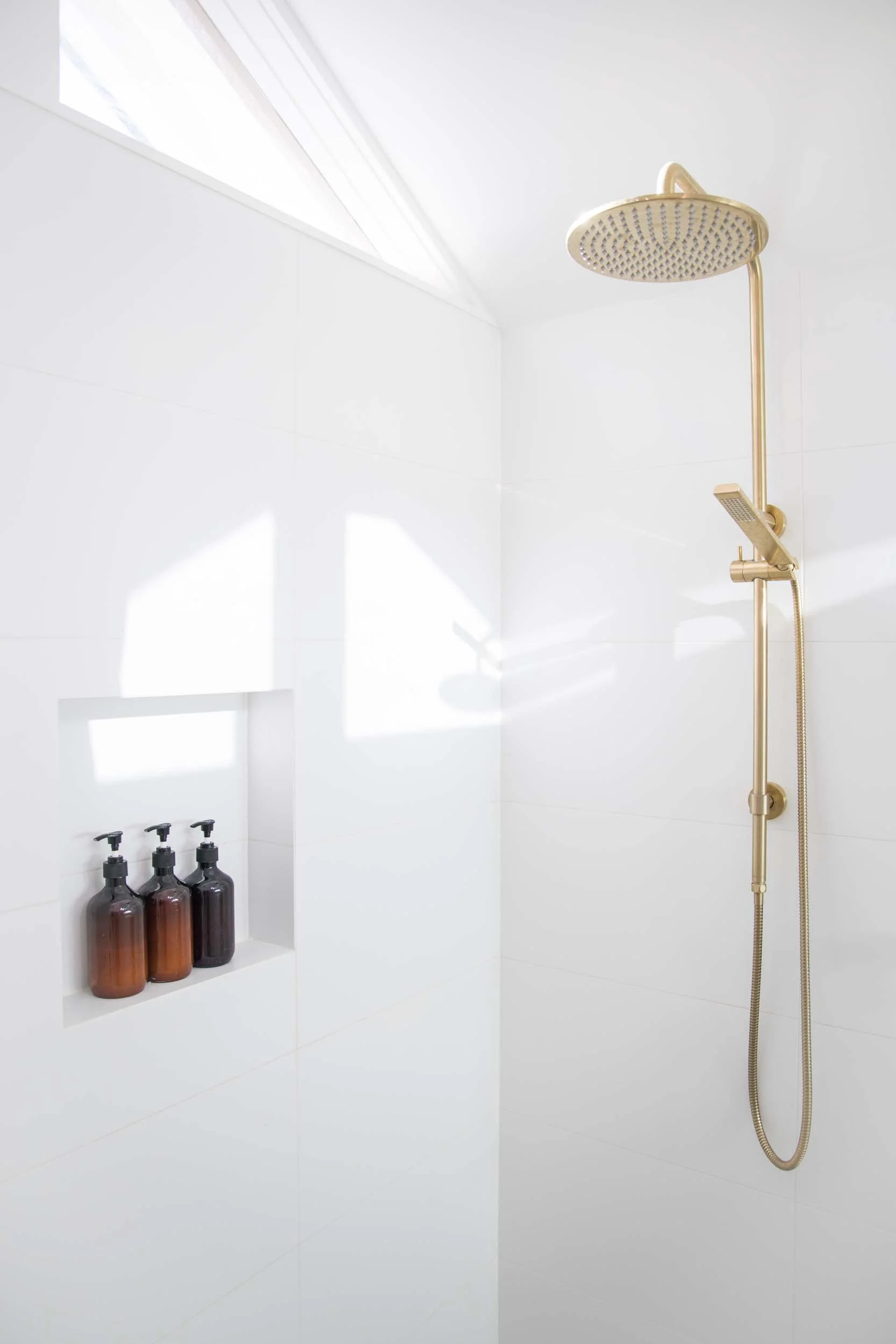 White shower with a round overhead showerhead, a handheld shower, and three bottles of soap or shampoo in a built-in niche.