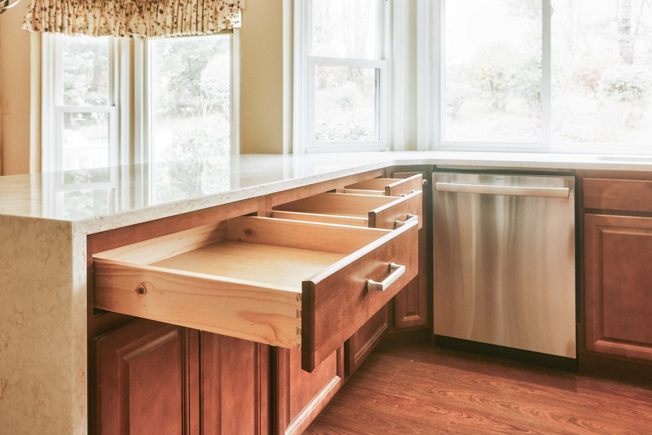 Empty kitchen cabinets with wooden drawers, a marble countertop, a stainless steel dishwasher, and a window with curtains letting in sunlight.
