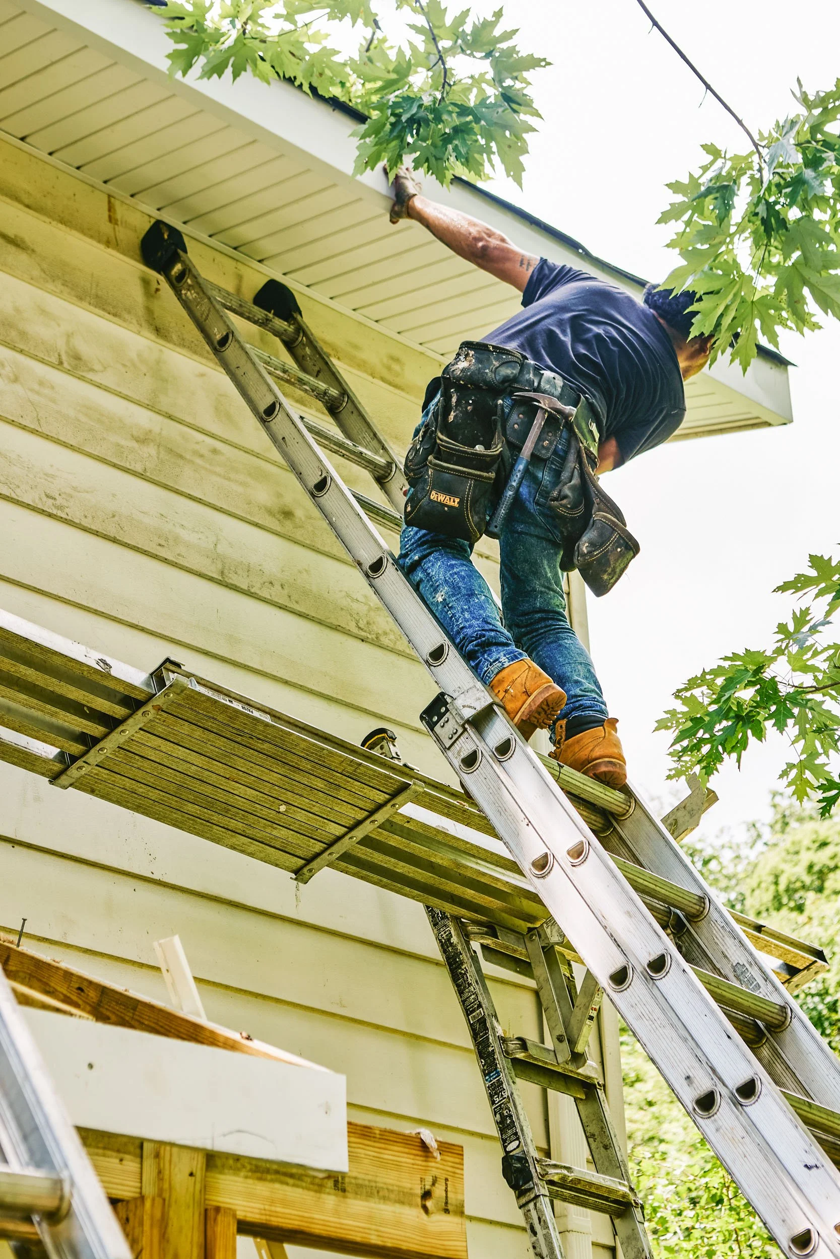A professional wearing work boots and a tool belt is installing new gutters on the exterior of a house, standing on a ladder and reaching up to the roof. Green foliage is visible overhead.