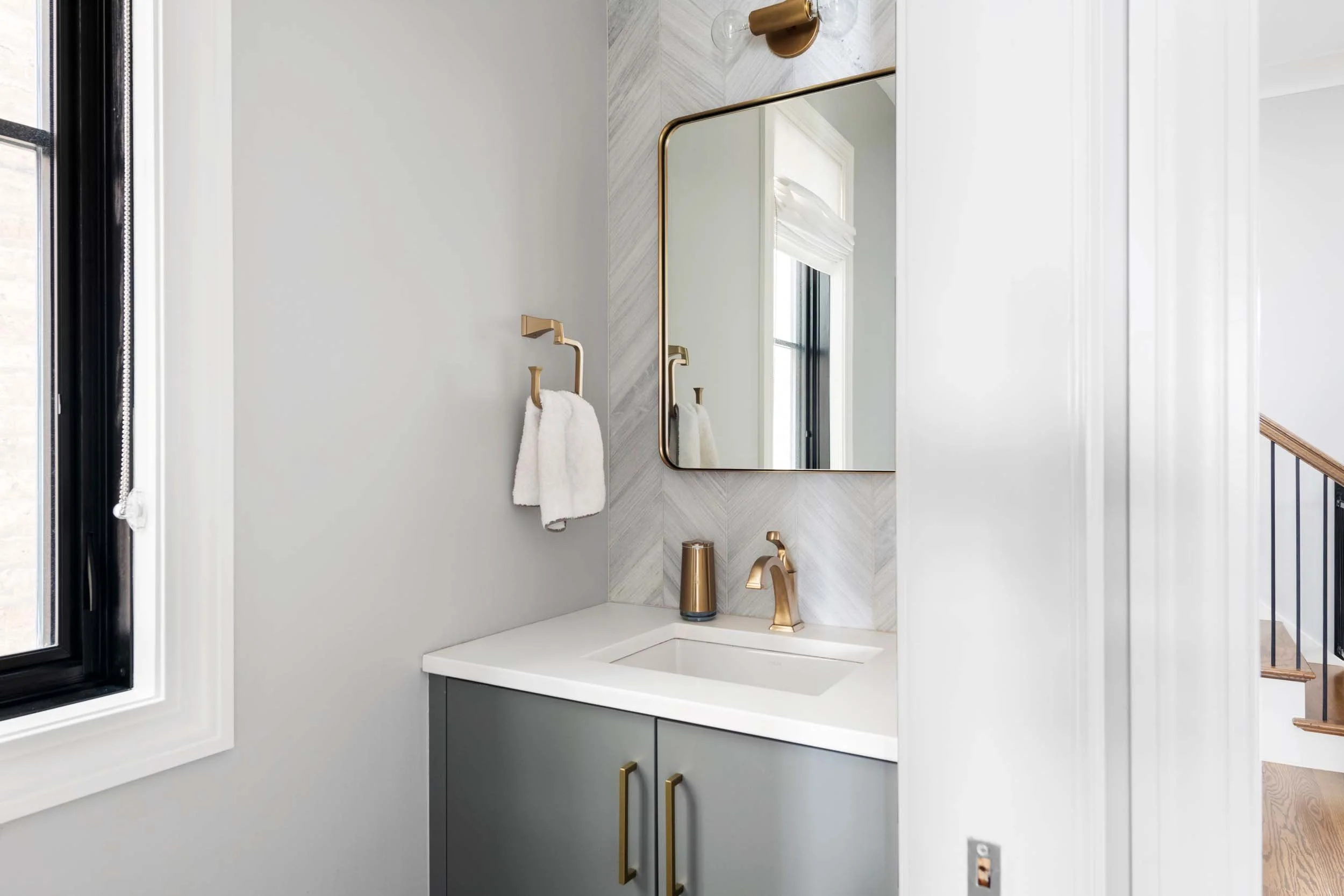 Modern bathroom vanity with gray cabinet, white countertop, and faucet, mirror with gold trim, wall-mounted towel ring with white towel, window with black frame, and staircase visible in the background.