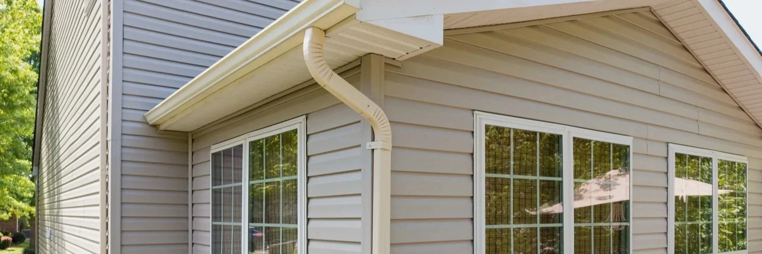 Close-up of a beige house exterior with vinyl siding, three windows, and a white gutter system.