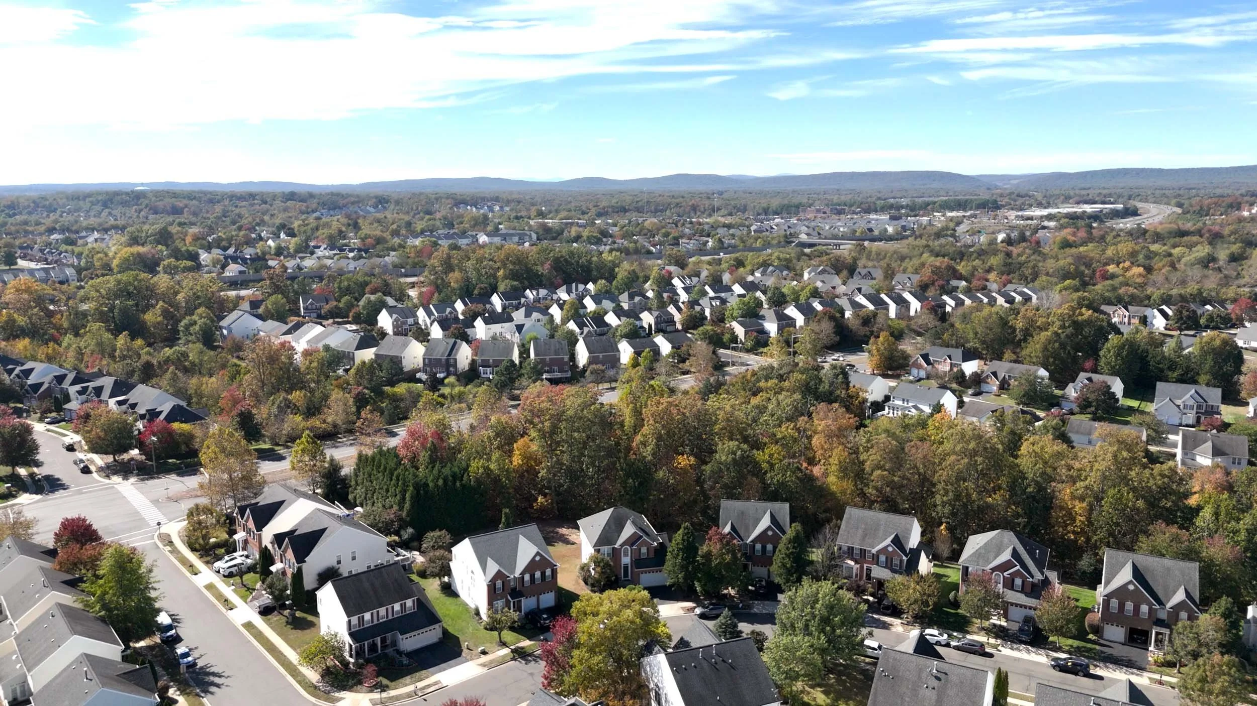 An aerial view of a suburban neighborhood with numerous houses surrounded by trees with changing autumn leaves. The area has well-maintained streets and a clear blue sky overhead.