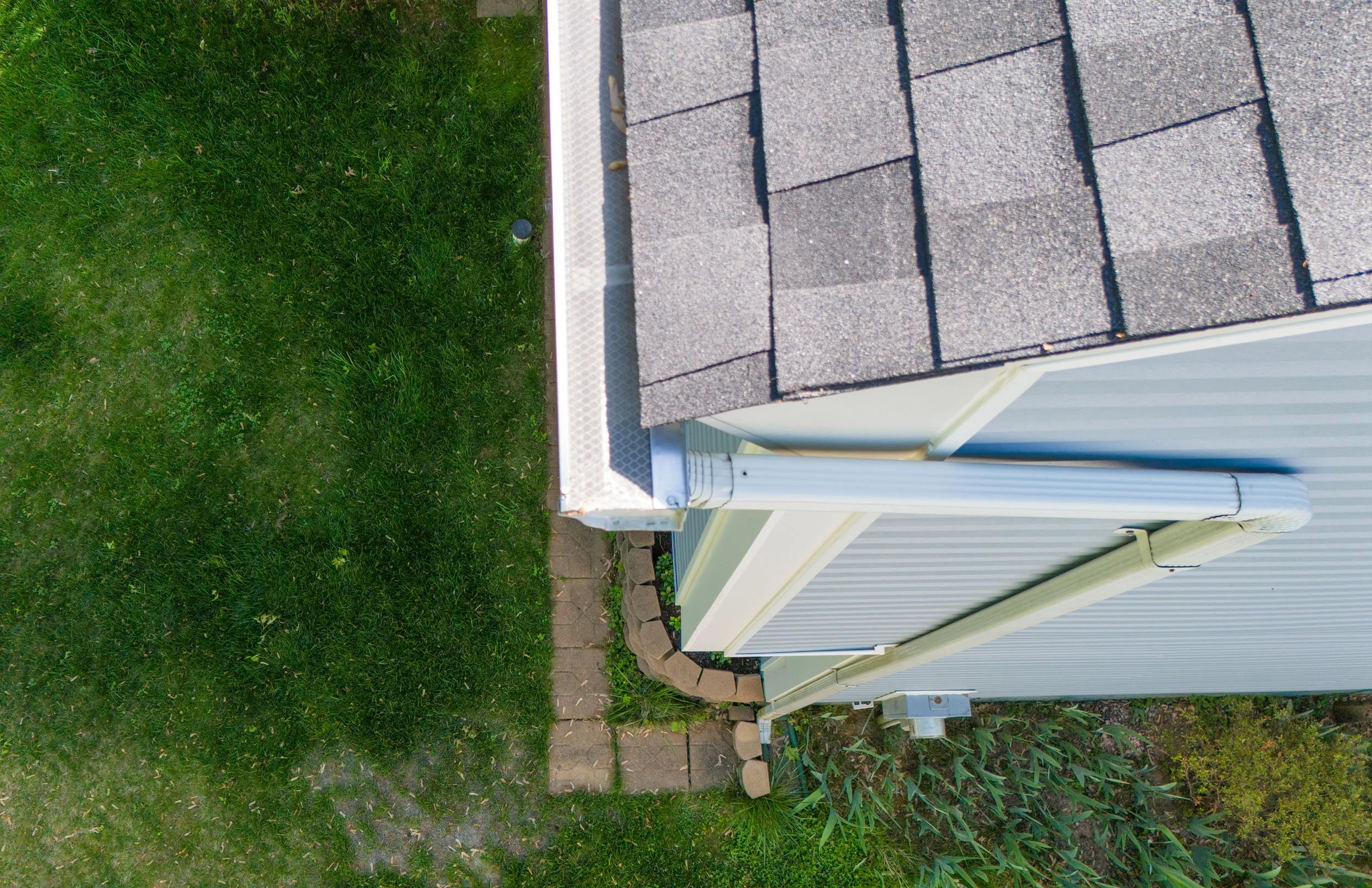 Upside-down view of the corner of a house with gray shingled roof, white exterior wall, and white gutter, next to a green lawn with small paver pathway and plants.