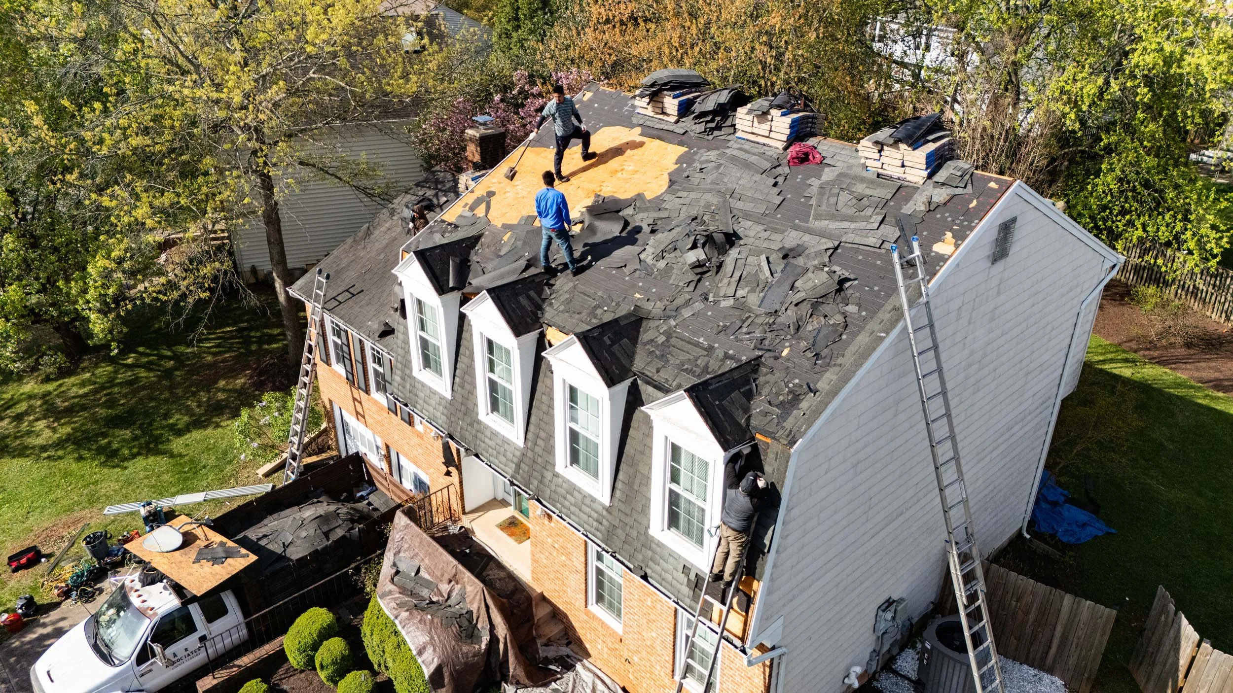 Aerial view of a house roof under renovation with workers removing old shingles, ladders on both sides, and construction materials around.