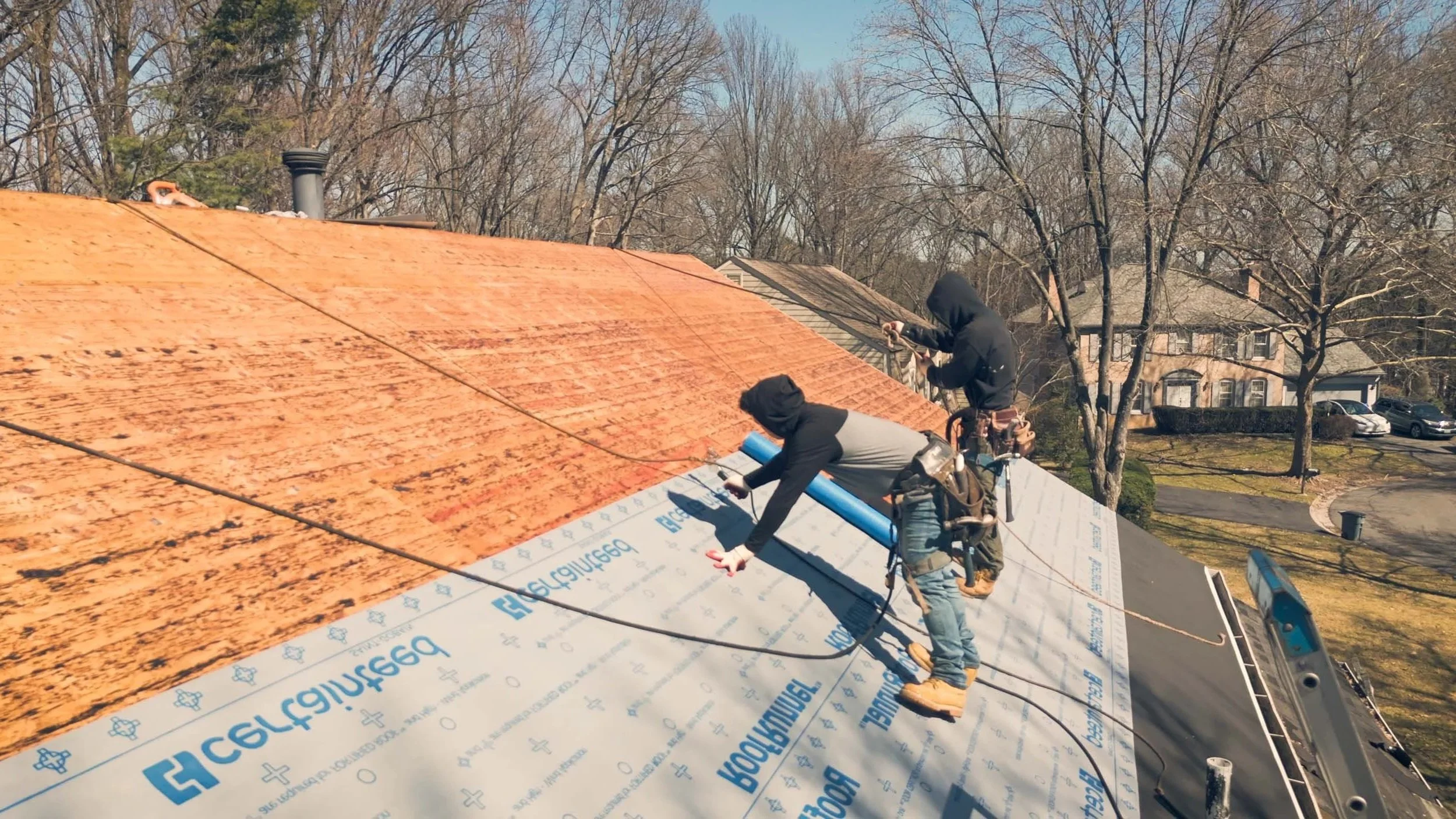 Two workers on a roof installing new roofing shingles, with houses and trees in the background.