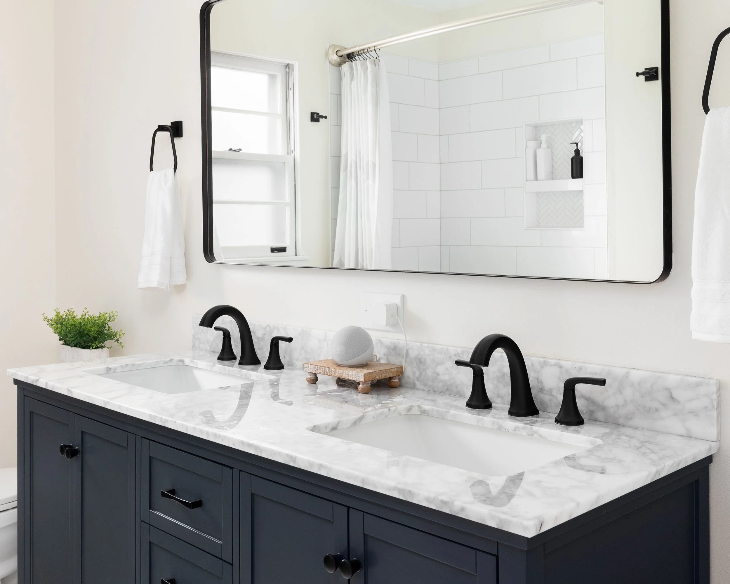 Bathroom double vanity with white marble countertop, black faucets, large mirror, white towels, a small plant, and a view of the shower area with white tiled wall and built-in shelf