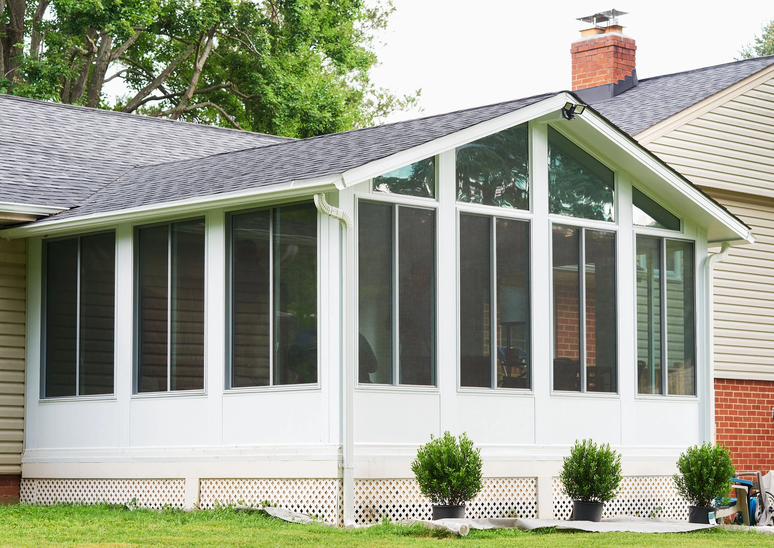 A house with a large enclosed porch featuring multiple windows, white framing, and a lattice skirting at the base. There are three potted bushes in front of the porch, a grassy yard, and trees in the background.