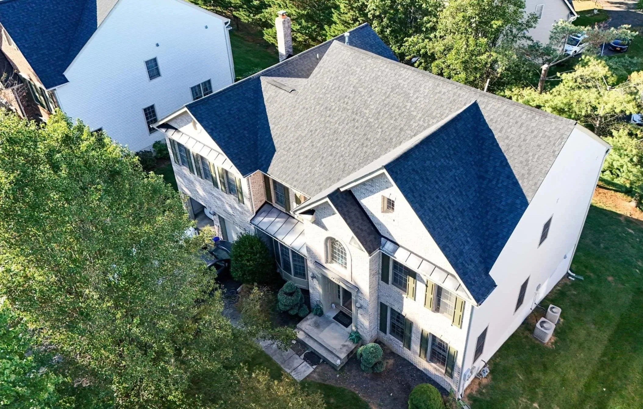 An aerial view of a large, multi-story house with a complex roof structure, surrounded by trees and neighboring houses in a residential neighborhood.