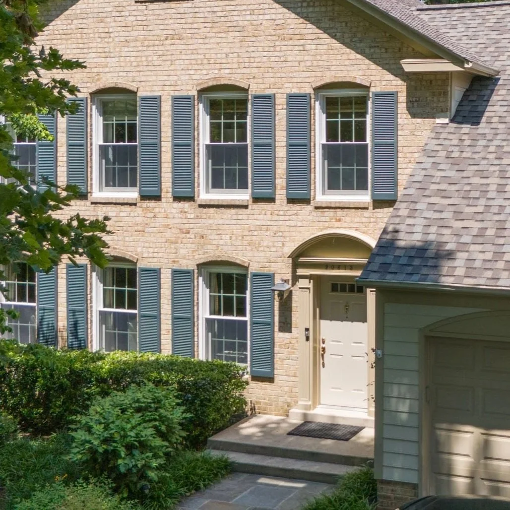 Front view of a two-story house with beige brick exterior, blue window shutters, and a white front door with a small porch.
