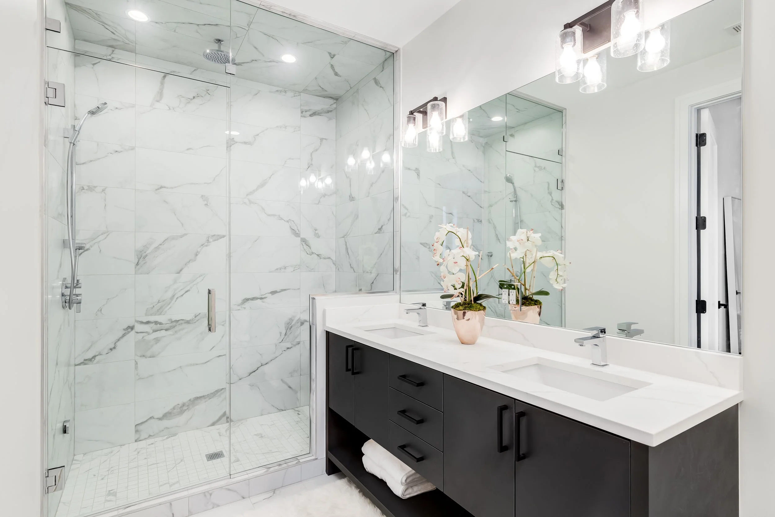 Modern bathroom with white marble shower and black vanity with double sinks, large mirror, potted orchids, and contemporary light fixtures.
