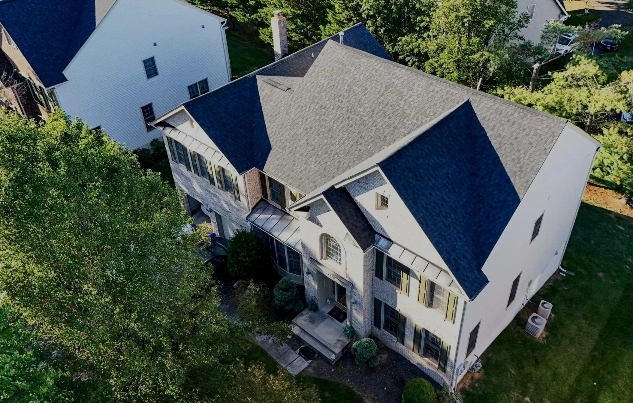 An aerial view of a large house with a dark gray shingled roof, surrounded by green trees and neighboring houses with white exteriors.
