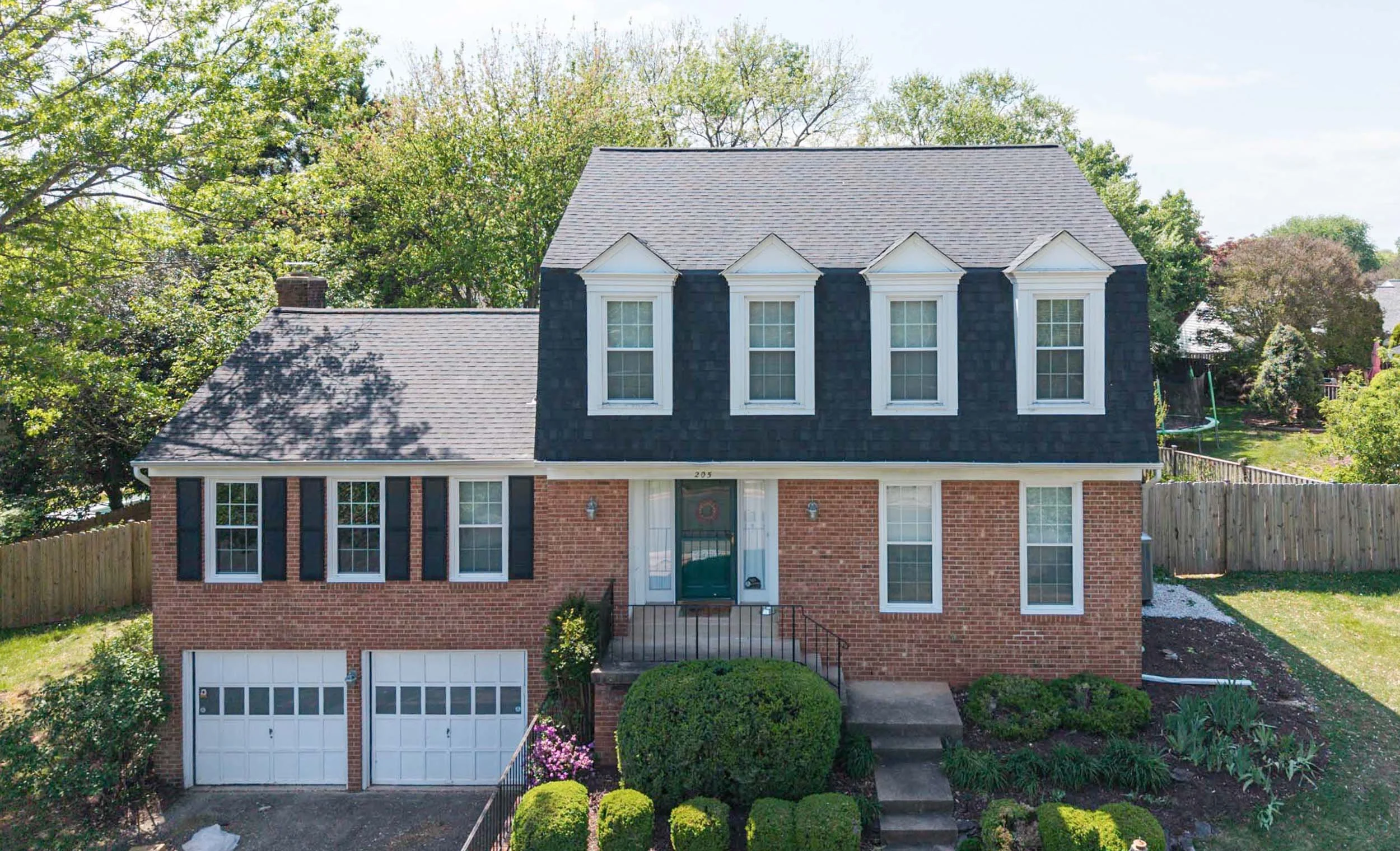 Front view of a two-story brick house with black shingle-style upper walls and white trim, featuring three dormer windows. The house has two cars parked in a double garage, a small front porch with black railings, and well-maintained landscaping with bushes and flowers. A wooden fence surrounds the backyard, and tall trees are in the background.