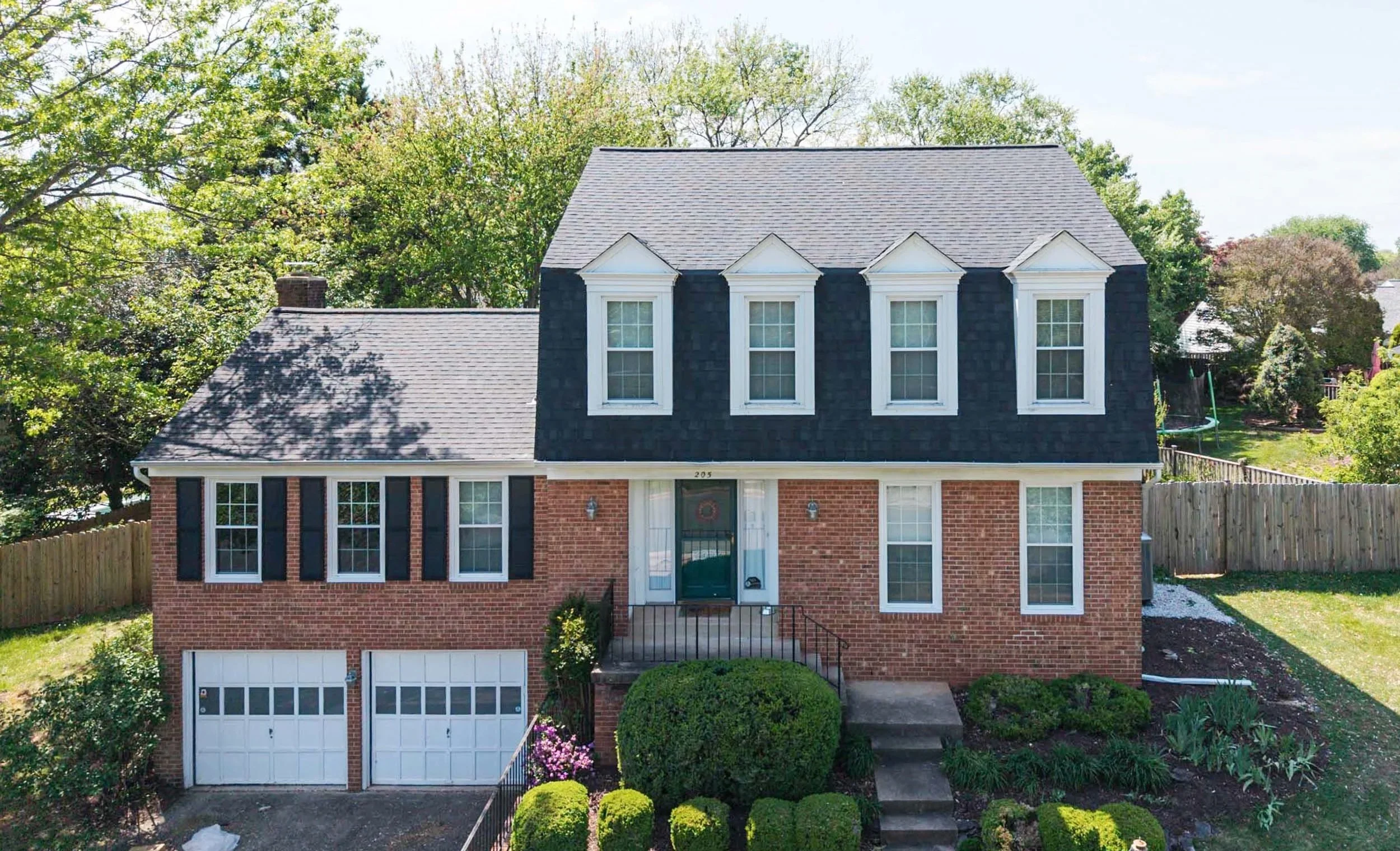 A two-story brick house with black shutters and a dark gray roof. There are five tall windows on the upper floor with white trim and decorative white pediments. The front door has a wreath and two small exterior lights on either side. The house has a lawn with bushes, and the driveway has a double garage with white doors. Surrounding the house are trees and a wooden fence.