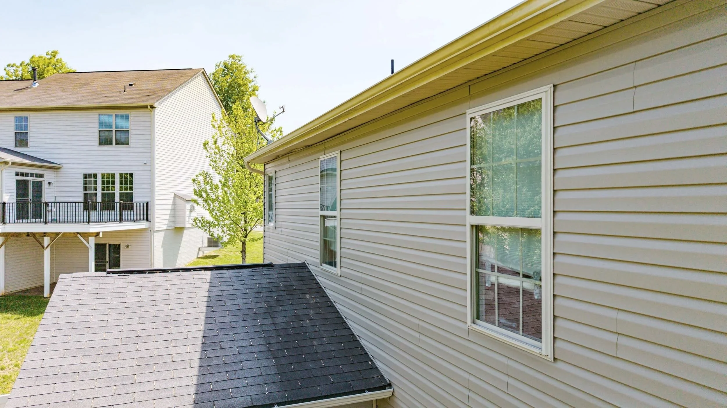 View of the side and roof of a beige house with vinyl siding, four windows, and a satellite dish, with a neighboring house and green trees in the background under a clear sky.
