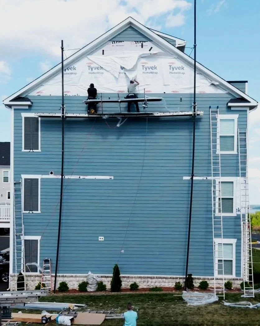 Construction workers installing siding on the upper part of a blue house, with ladders and scaffolding around the house.