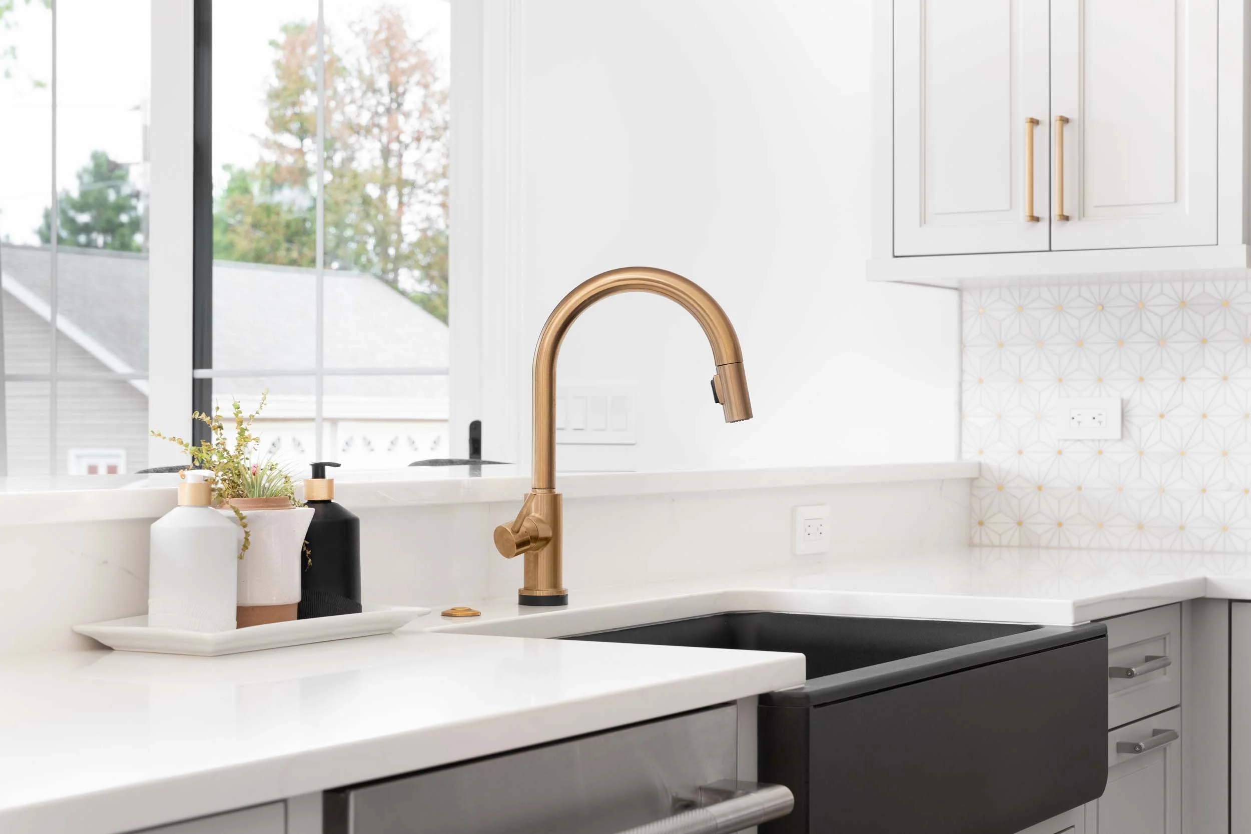 Modern kitchen sink with a gold faucet, white countertop, potted plants, and soap bottles, with a window showing an outdoor view.