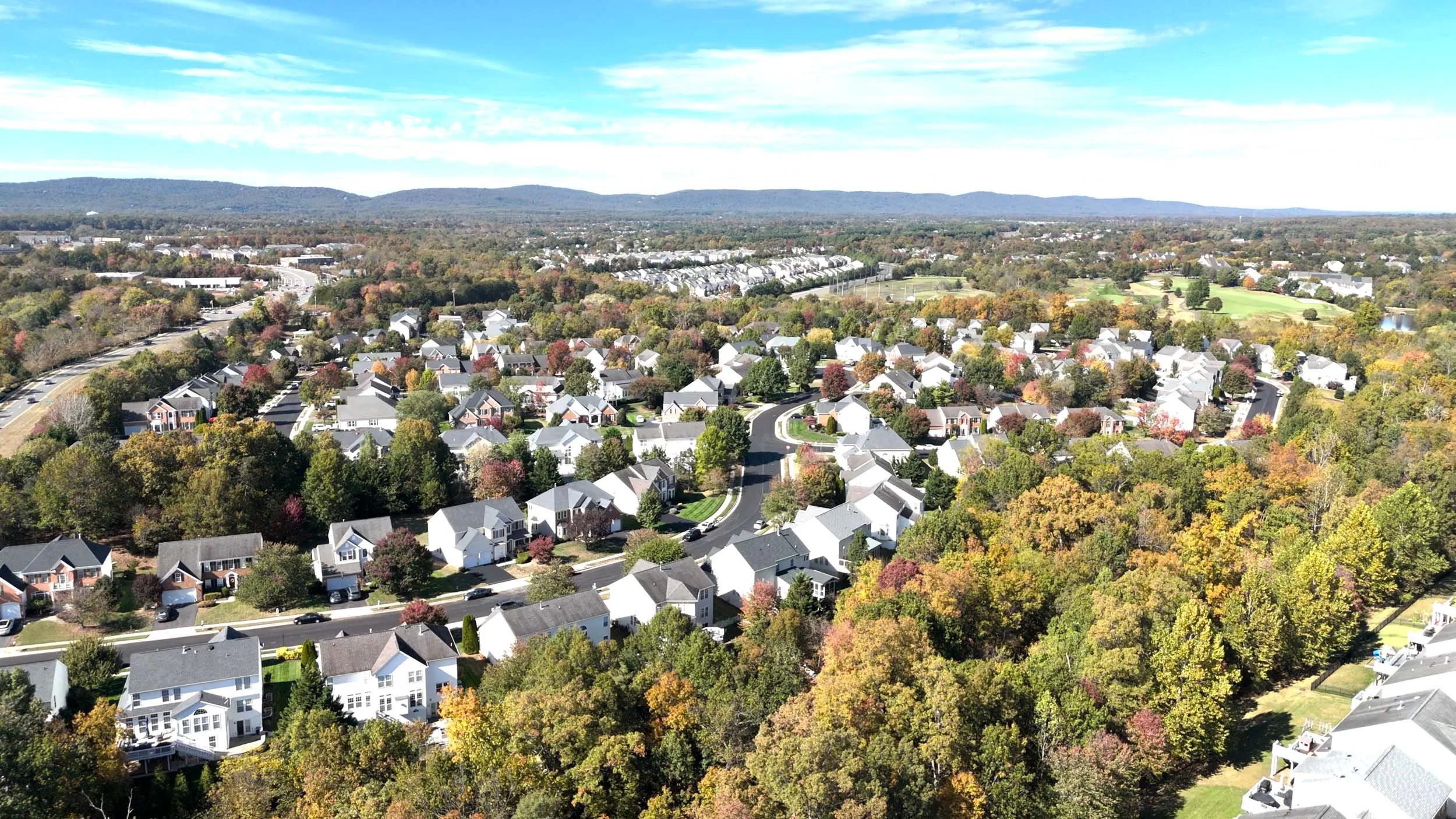 An aerial view of a suburban neighborhood with houses, trees, and roads, with hills and a partly cloudy sky in the background.