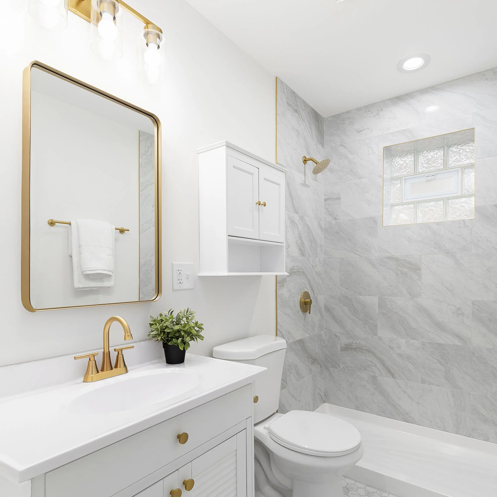 Modern white bathroom with gold accents, featuring a mirror, sink, toilet, plant, towel, and a walk-in shower with a textured gray marble wall and a window.
