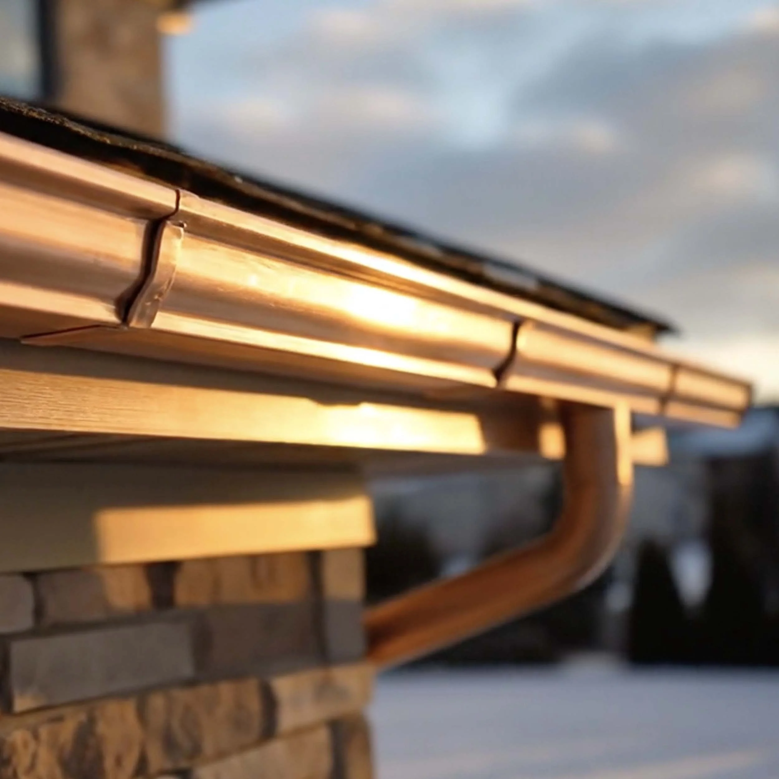 Close-up of a rooftop edge with golden sunlight, showing brown copper flashing and a wooden gutter pipe against a blurred sky background.