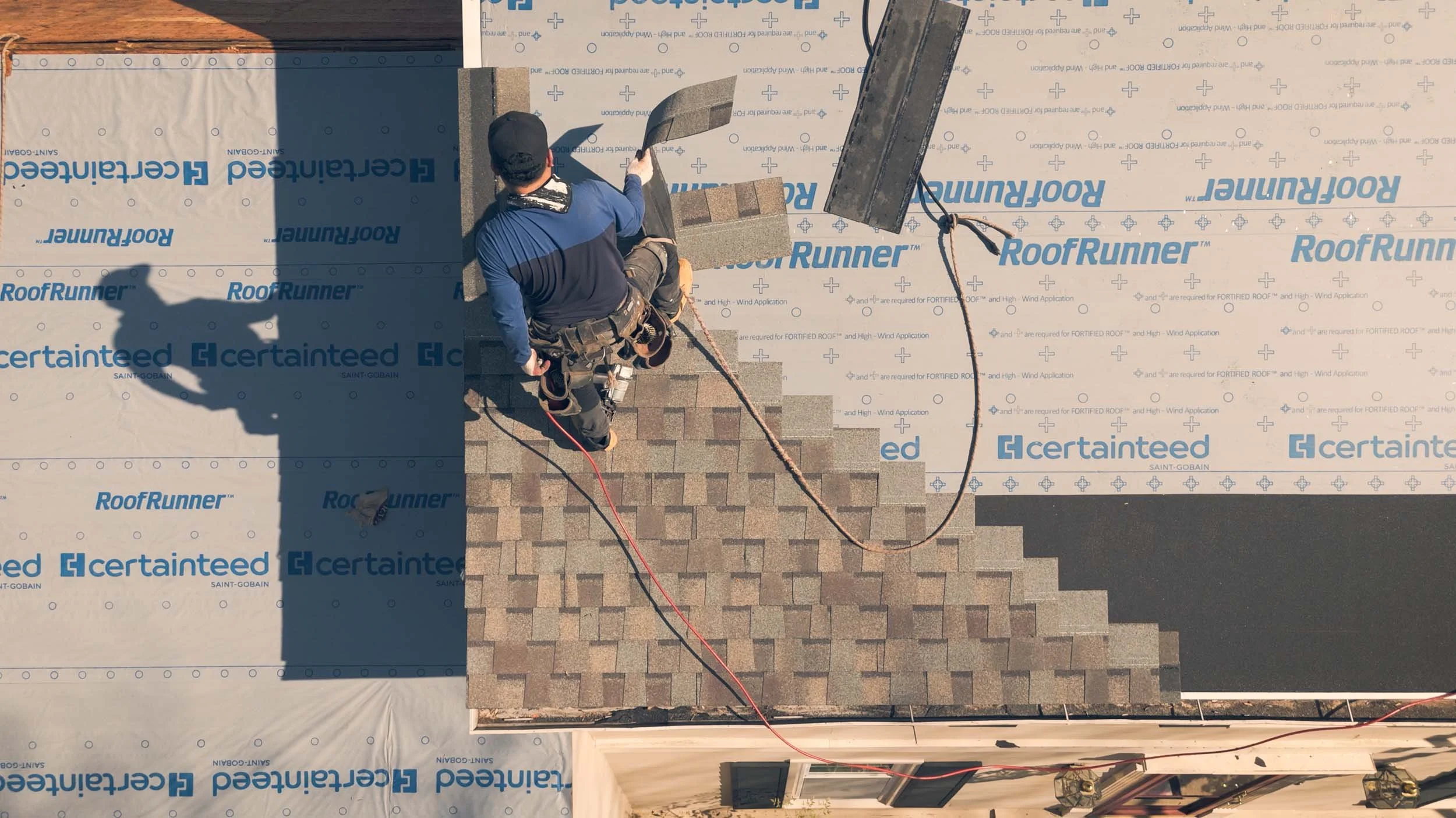 A worker installing shingles on a roof. The shingles are brown and there is roofing felt with blue and white branding underneath, indicating ongoing roofing work. The worker is wearing a black cap, a blue shirt, and a harness, and is working near the edge of the roof with some roofing tools and materials nearby.