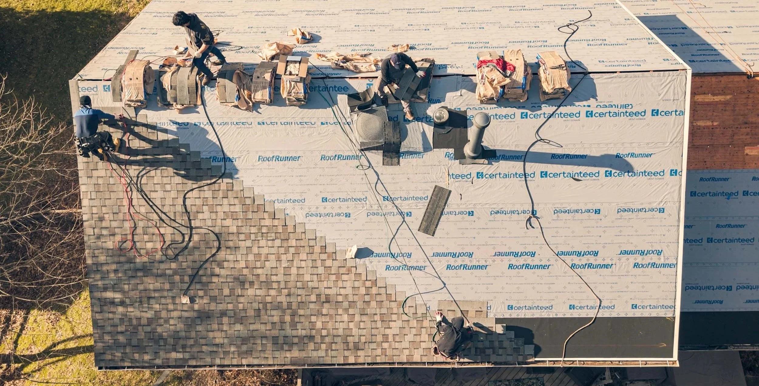 Aerial view of a rooftop under construction with workers installing roofing material, partially covered with a protective underlayment labeled 'CertainTeed' and 'RoofRunner', and an area with shingles already installed.