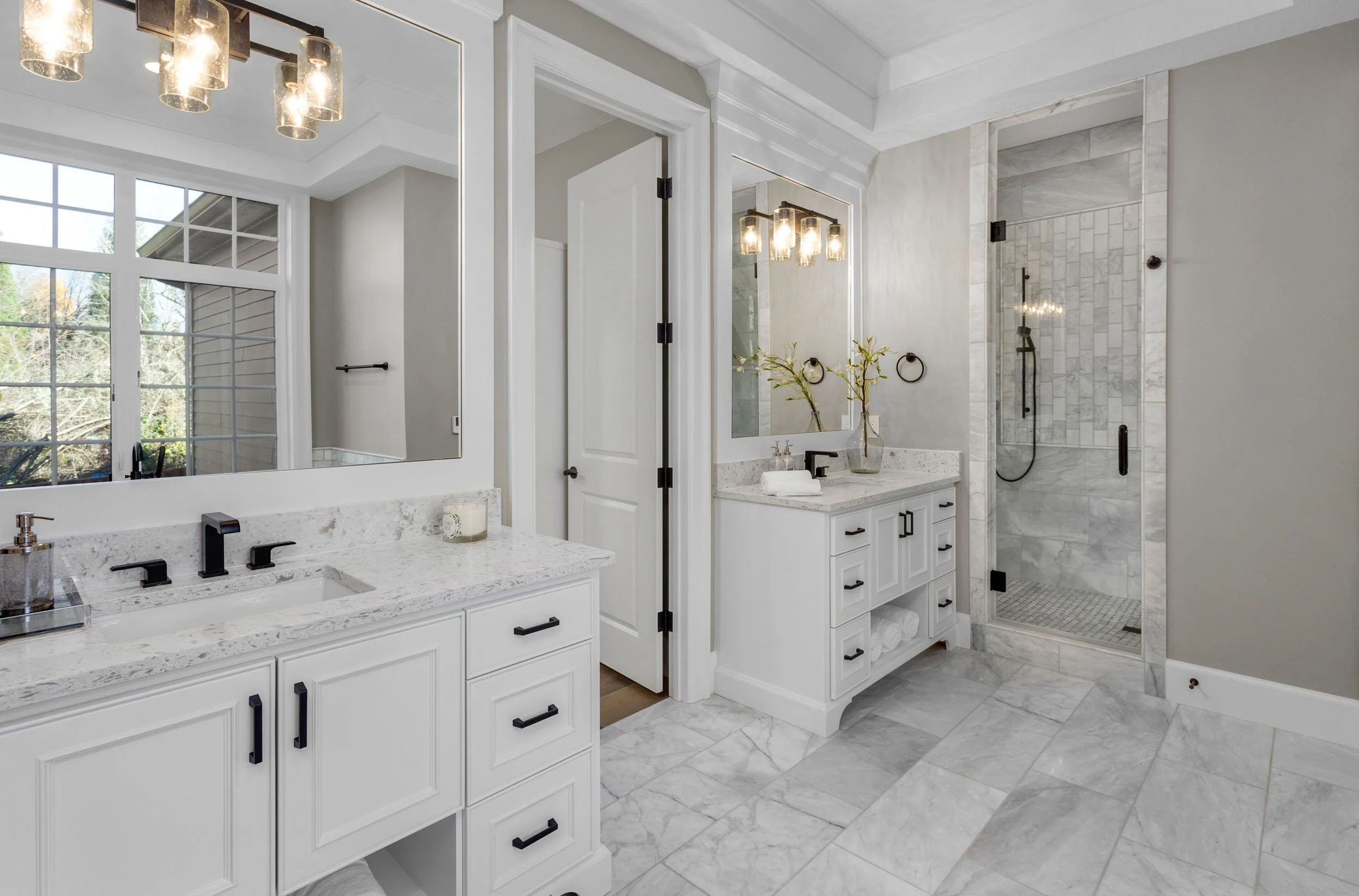 A modern, bright bathroom with white cabinets, marble countertops, and a walk-in shower with marble tiles and a glass door.