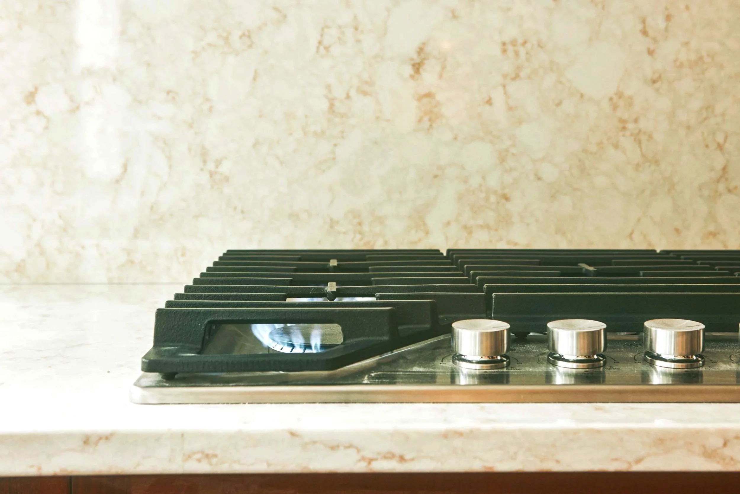 Close-up of a modern gas stovetop with three burners on a marble countertop, beige textured wall in background.