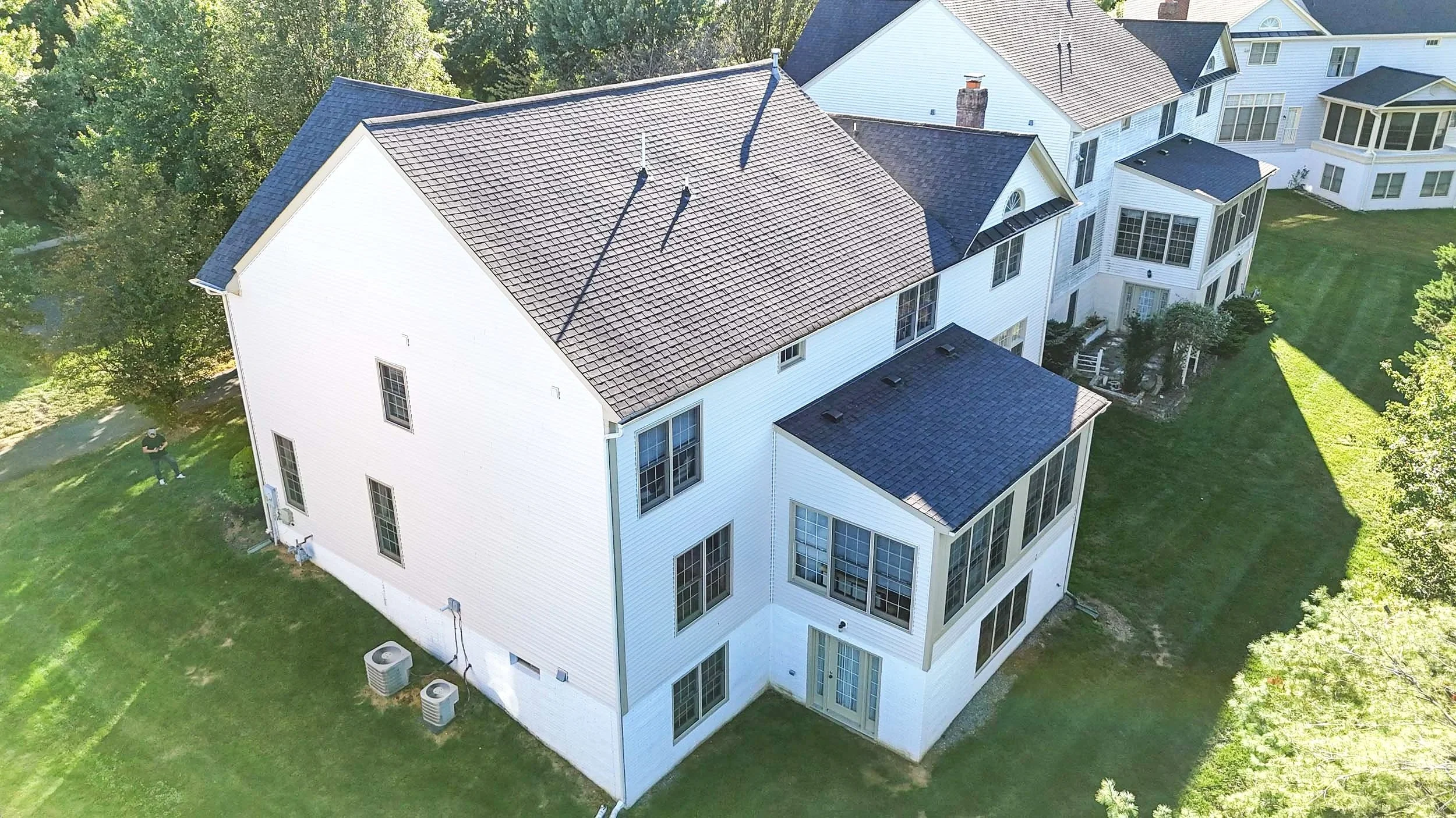 Aerial view of a white multi-story house with a pitched roof, multiple windows, a small patio area, surrounded by a lawn and trees.