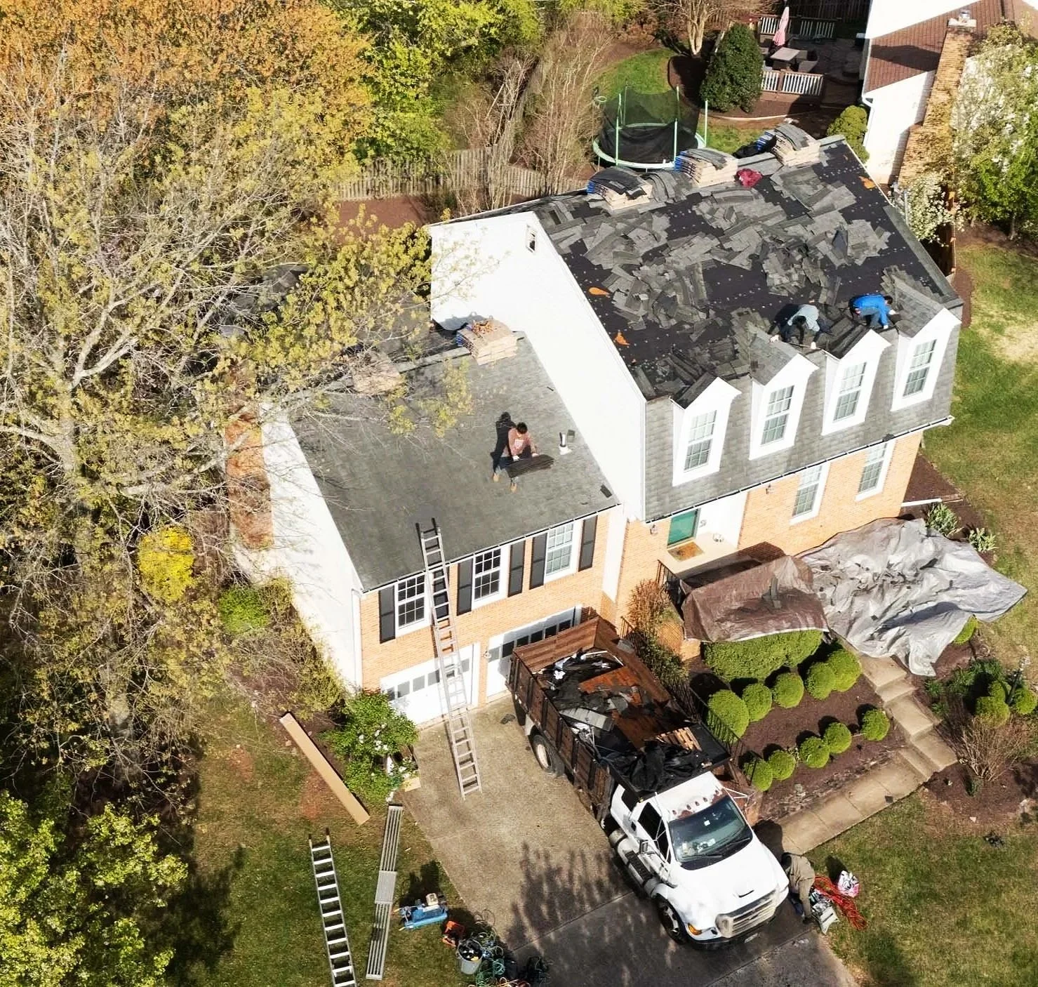An aerial view of a two-story house where roof repair work is in progress. Two workers are on the roof, replacing or fixing shingles. Several ladders are leaning against the house, and a truck with roofing supplies is parked in the driveway. The backyard has a trampoline, a covered object, and trees with autumn foliage.