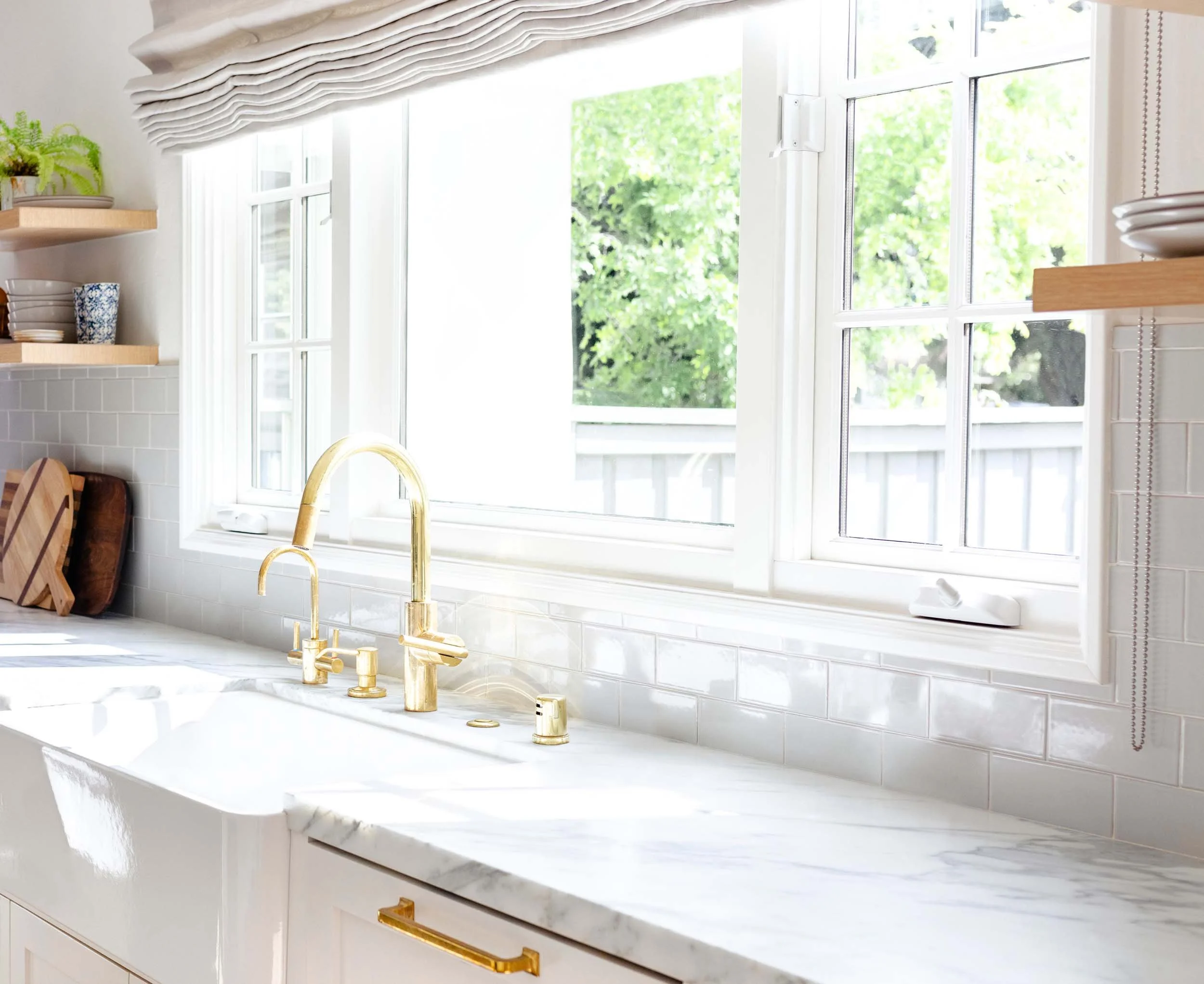 Bright kitchen with white cabinets, marble countertop, gold faucet, and open window showing green trees outside.