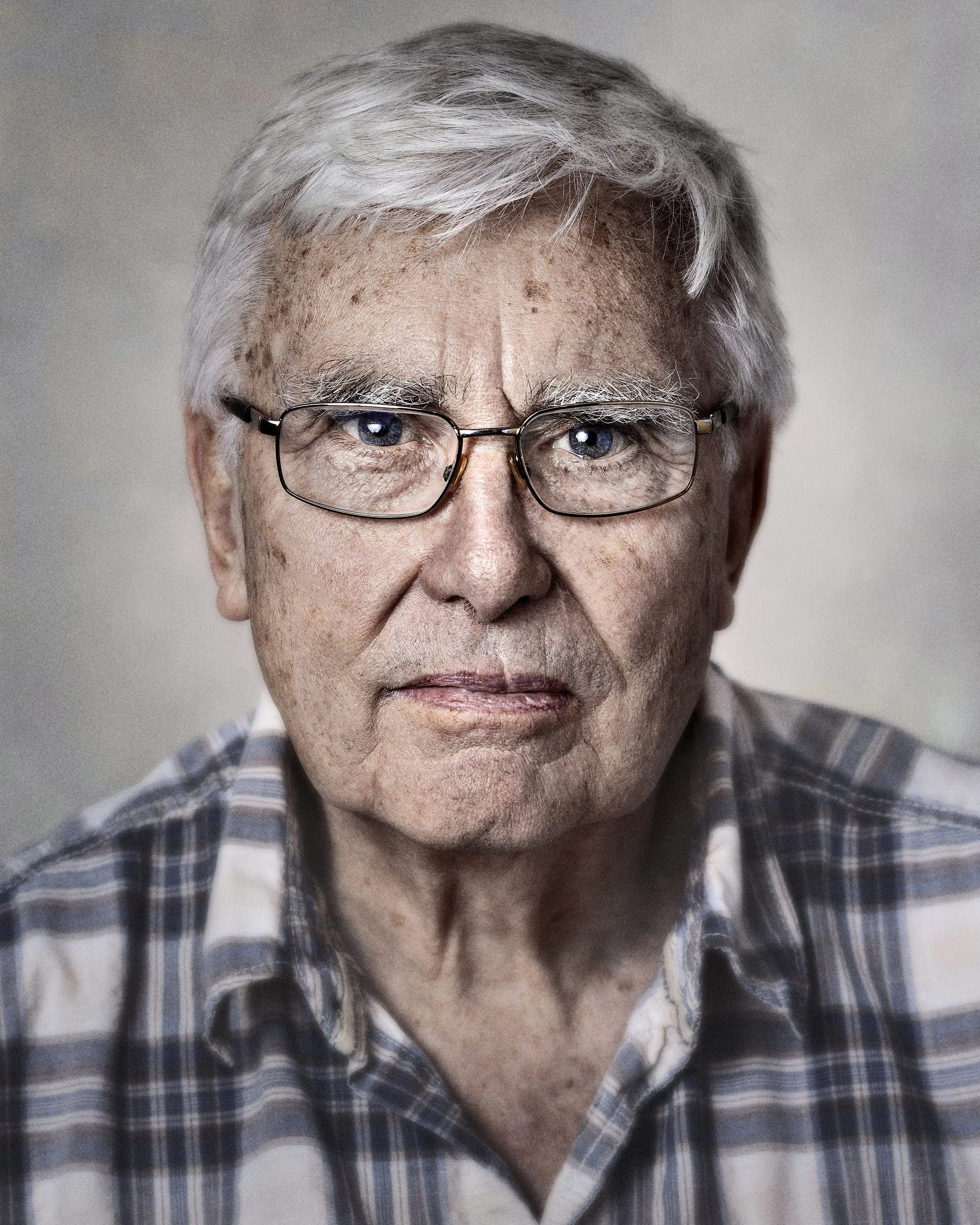 Close-up of an elderly man with gray hair and glasses, wearing a plaid shirt.