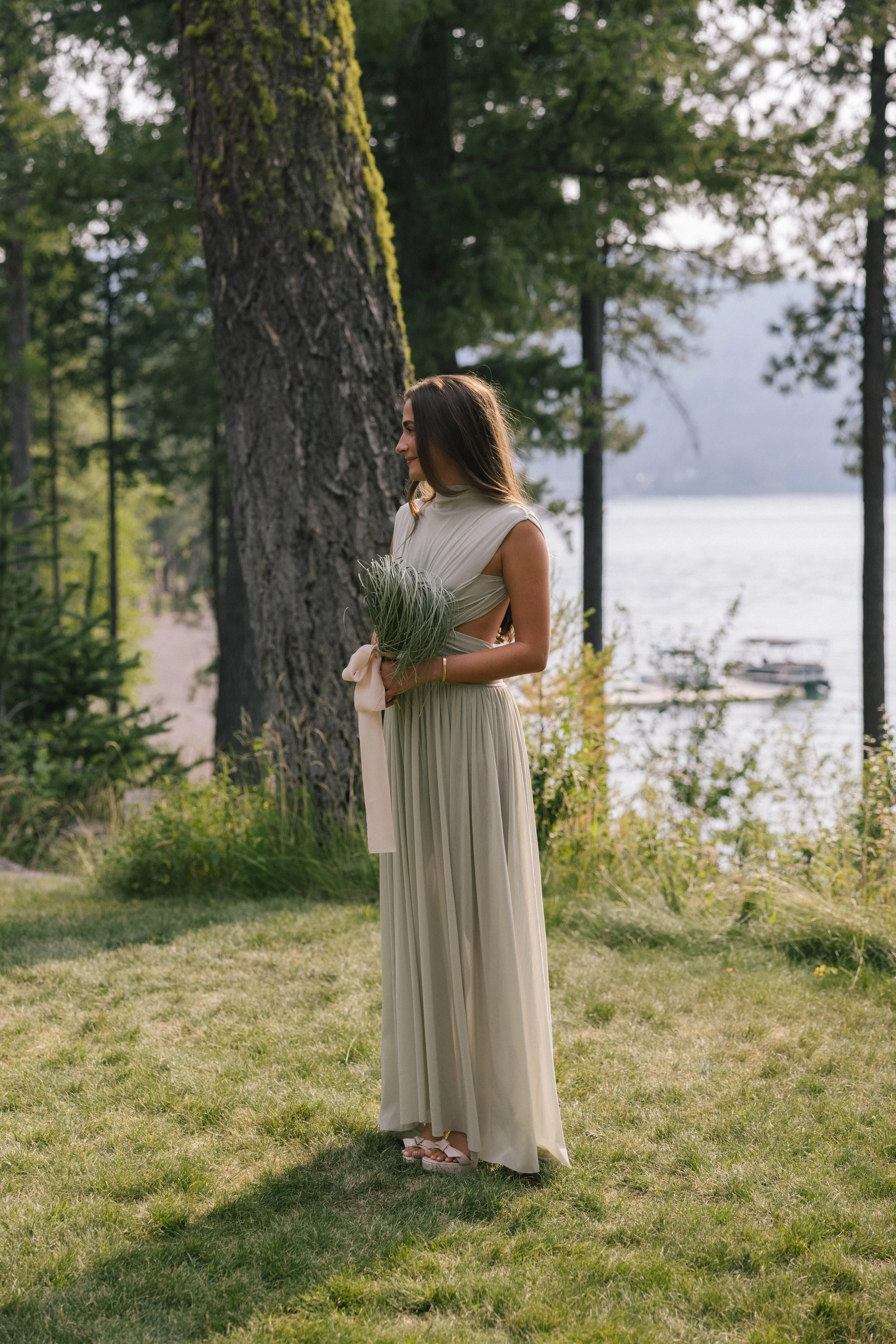 Woman in a light green dress holding a small bouquet of greenery, standing on grass near a lake with trees in the background.