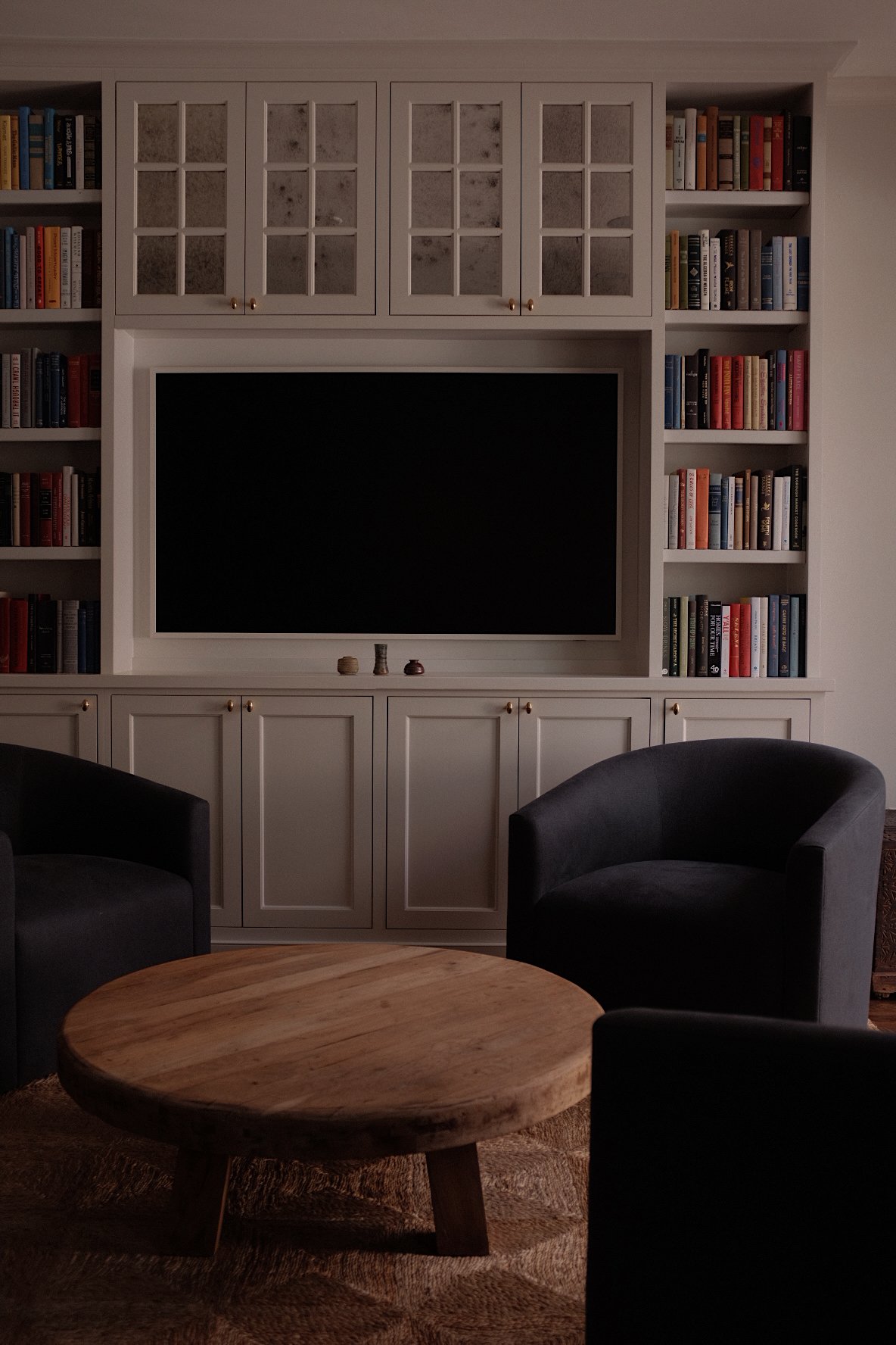 A living room with a large black television centered in a white built-in bookshelf and cabinet. The shelves are filled with colorful books. In front of the TV is a round wooden coffee table. Two dark gray armchairs are partially visible in the foreground.