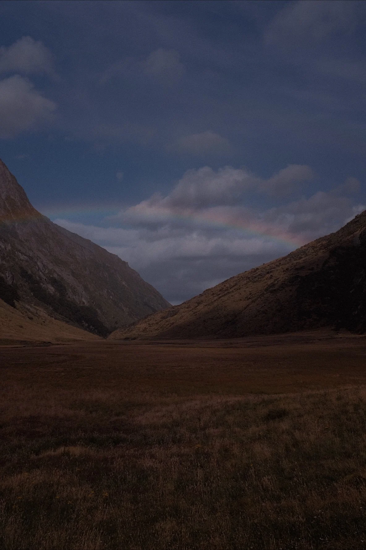 Landscape of a valley between two hills under a cloudy sky, with a faint rainbow in the background.
